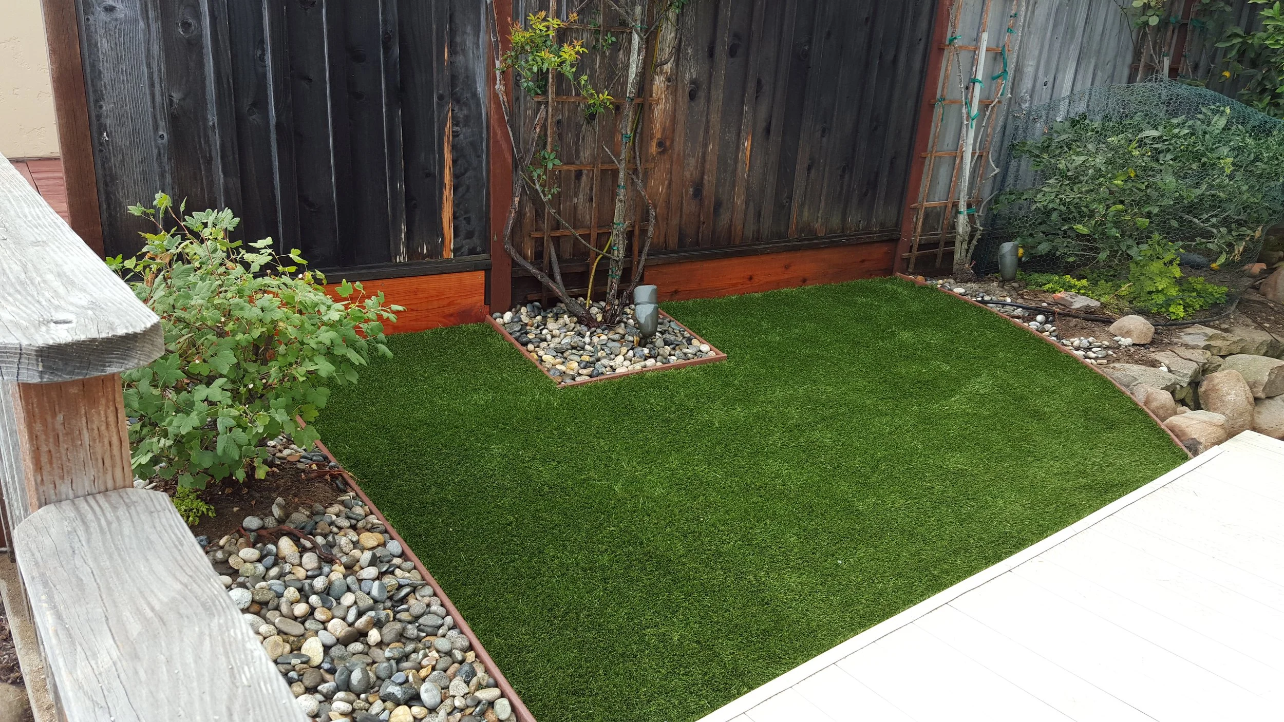 A small backyard with artificial grass, bordered by rocks, a wooden fence, and some plants.