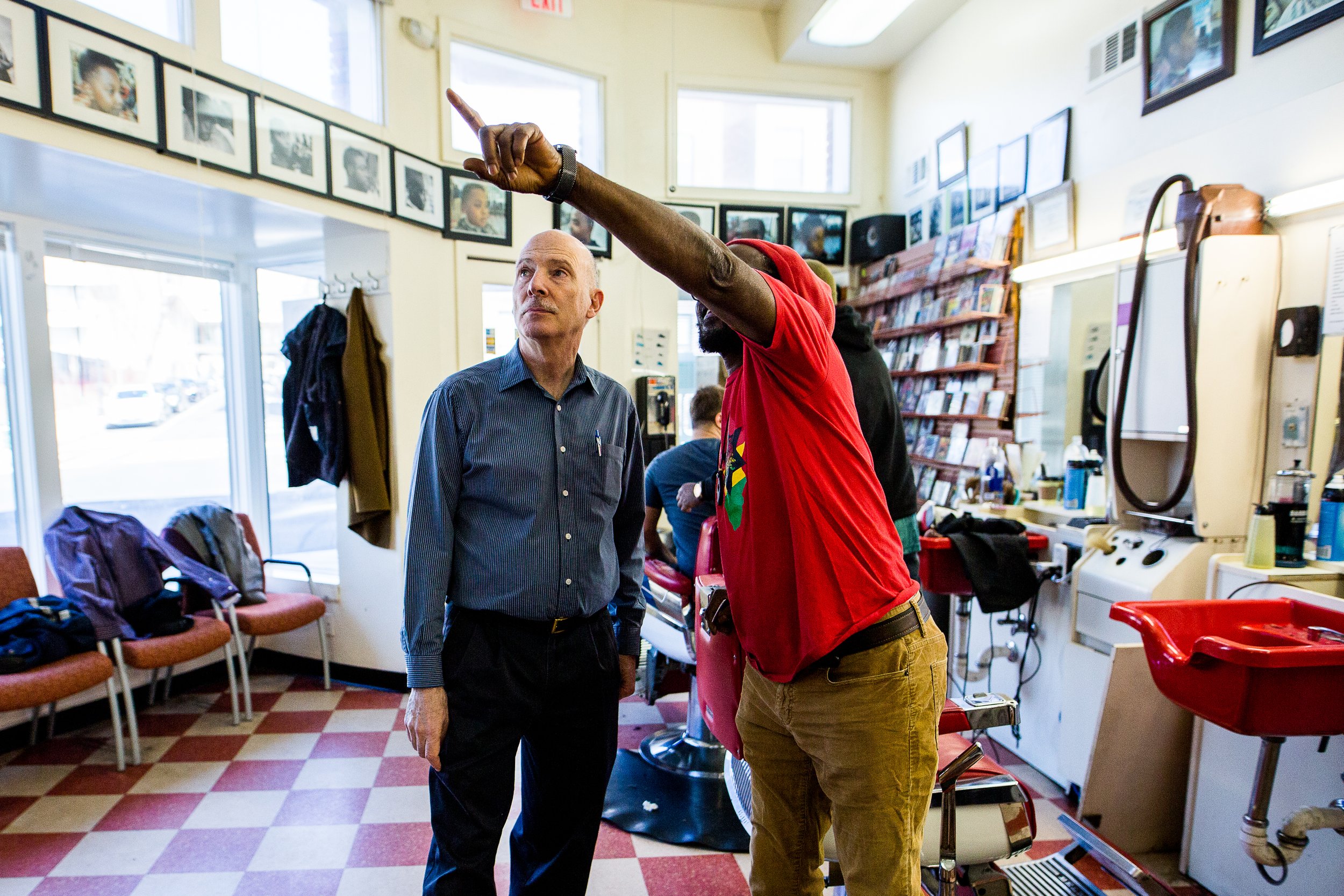 A bald man in a blue shirt and black pants standing next to a man in a red t-shirt and brown pants in a barbershop or salon, pointing with his right hand. There are chairs with backpacks and jackets, a window, and shelves with photos and magazines on the walls.