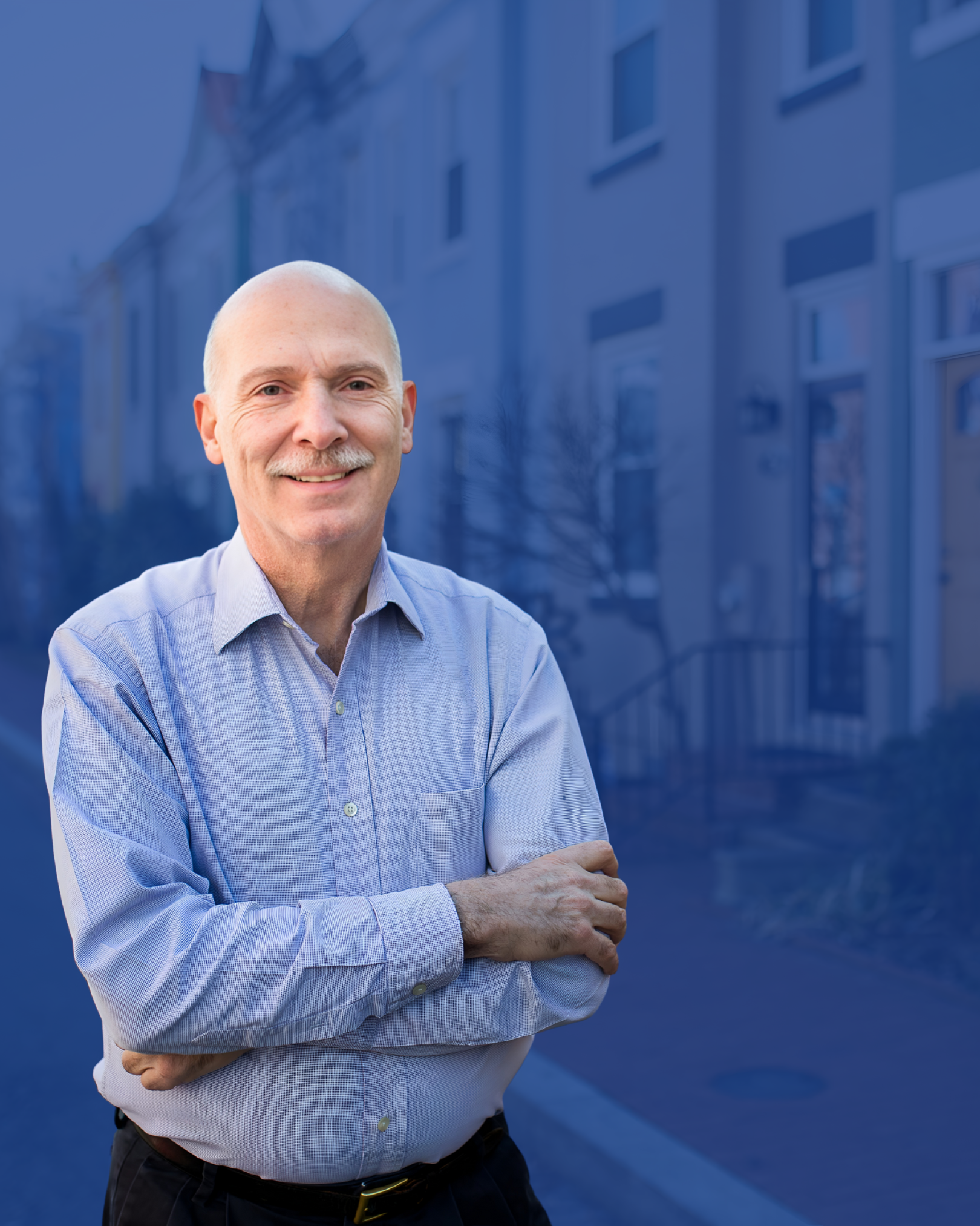 A smiling bald man wearing a light blue button-up shirt standing outdoors with arms crossed, in front of a blue-tinted background of buildings and trees.
