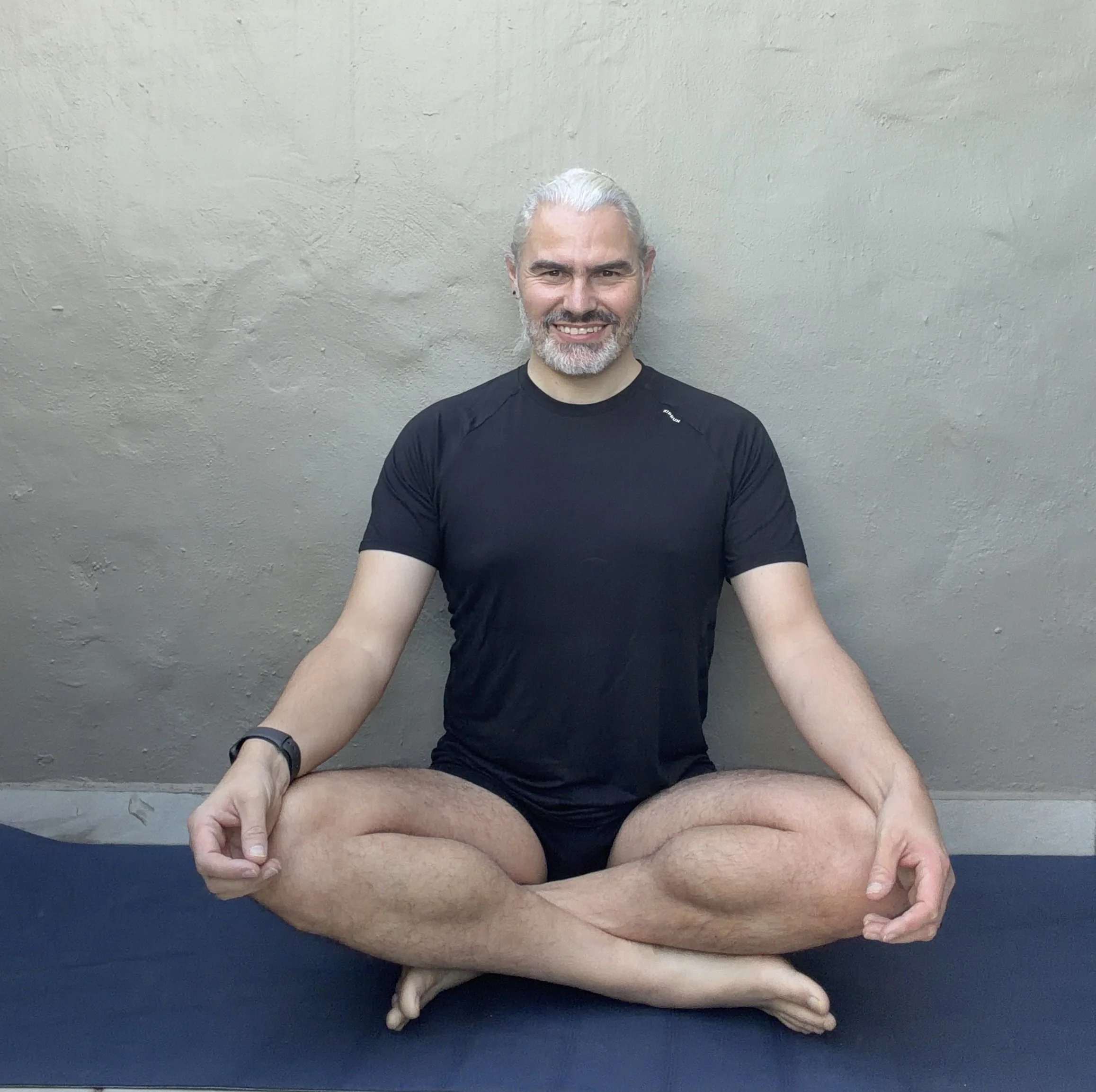 A man with white hair and beard sitting cross-legged on a yoga mat against a plain wall, wearing a black t-shirt and shorts, smiling.