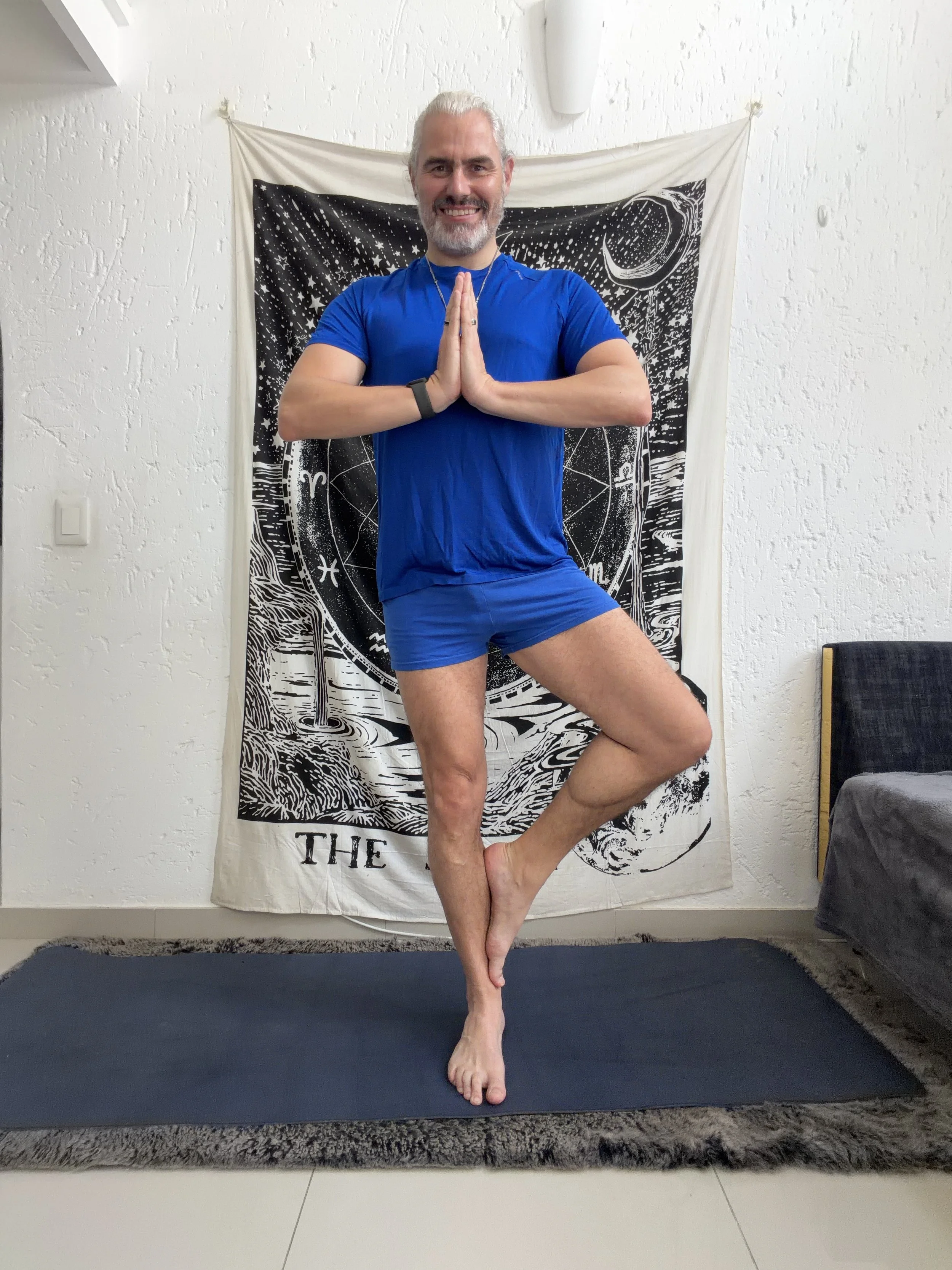 A man practicing yoga indoors on a yoga mat, standing in a tree pose with hands in prayer position and one foot resting on his inner thigh. He is smiling and wearing a blue athletic shirt and shorts, with a tapestry featuring celestial designs hanging on the wall behind him.