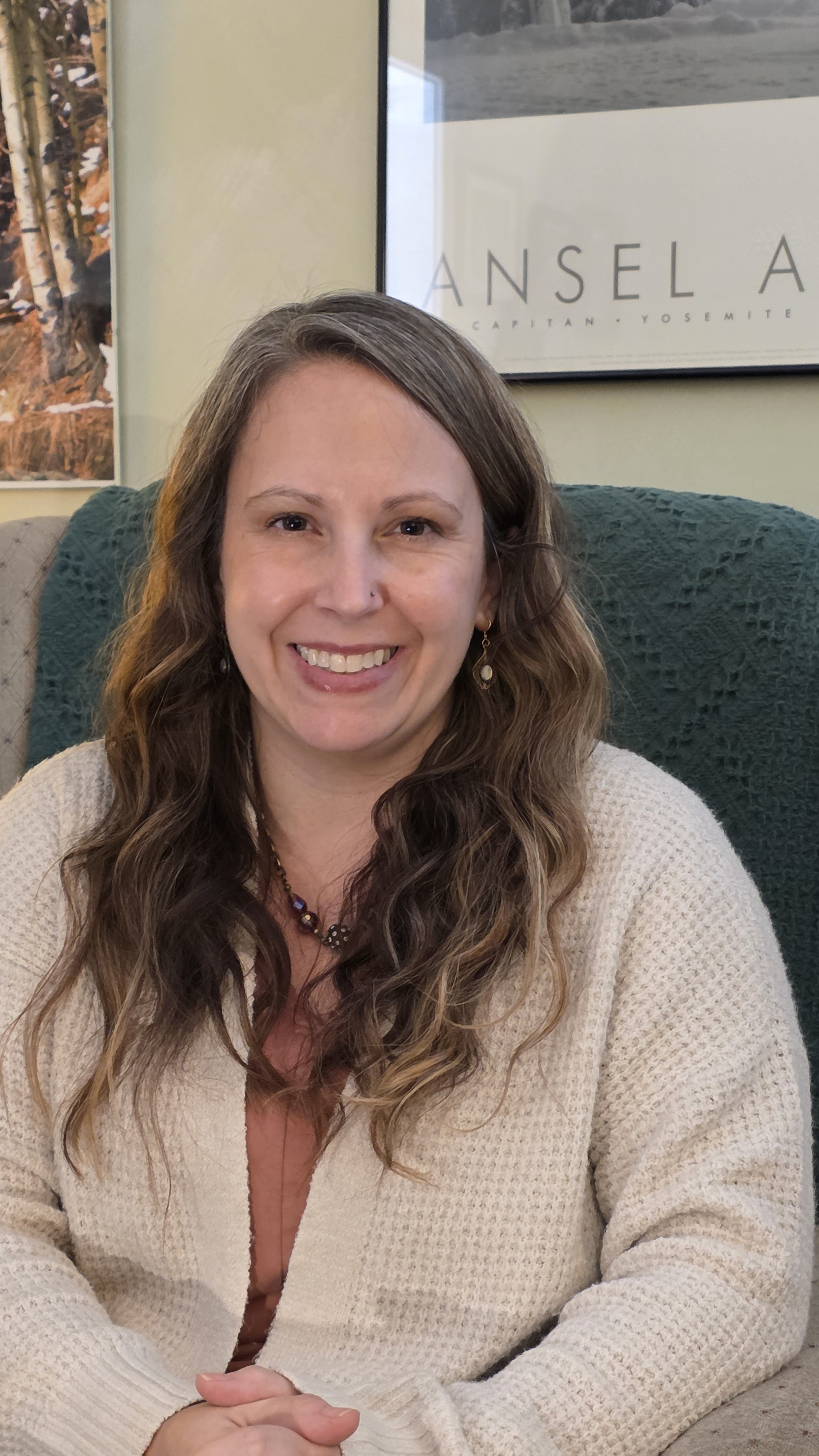 A smiling woman with long, wavy brown hair, wearing earrings and a beige sweater, sitting on a couch with a green blanket. Behind her, there are framed pictures on the wall, including one with text that says "Ansel A" and features mountain scenery.