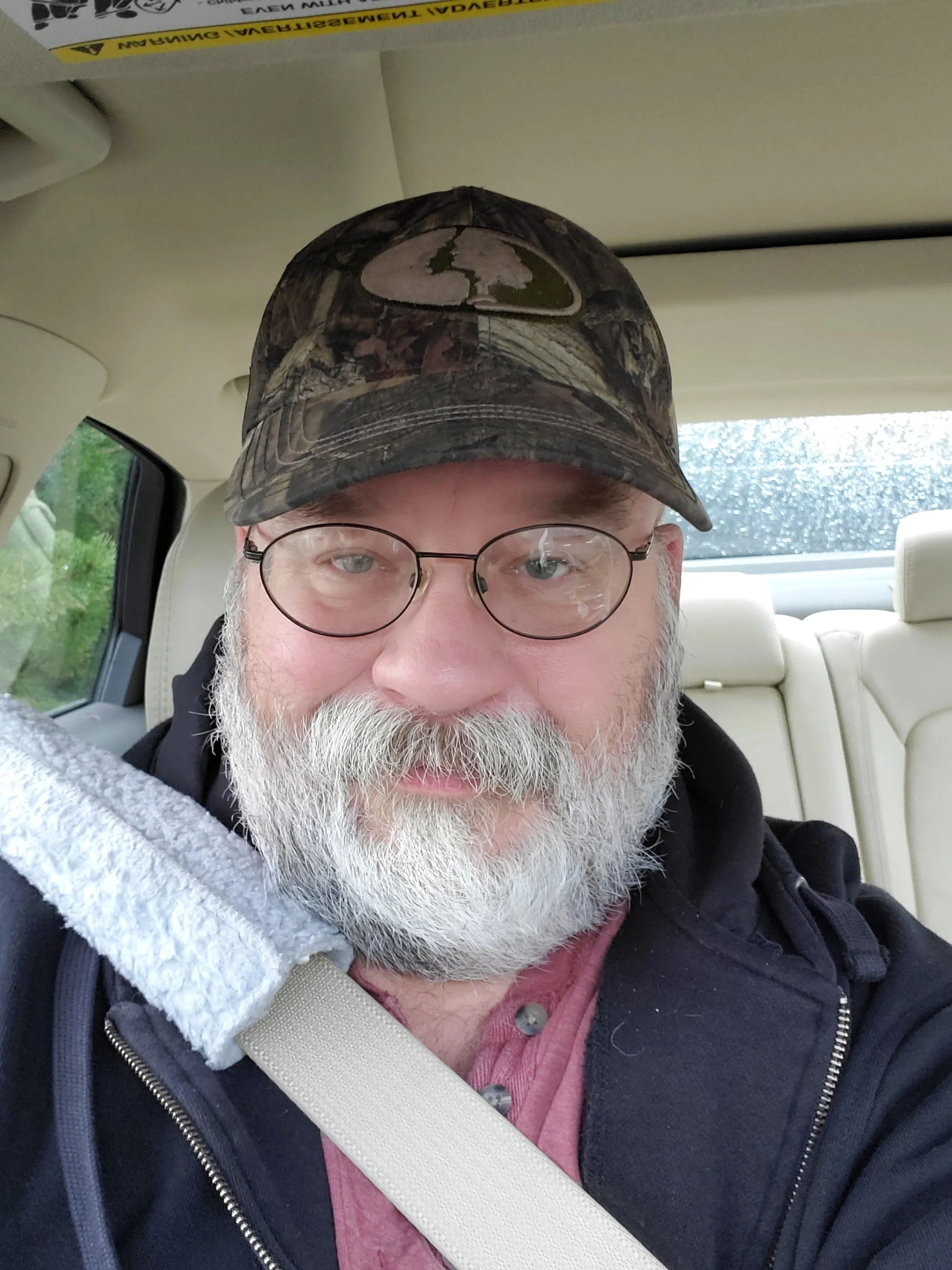 Close-up of an older man with a grey beard and glasses, wearing a camouflage cap, a red shirt, and a black hoodie, seated in a vehicle with a beige interior.
