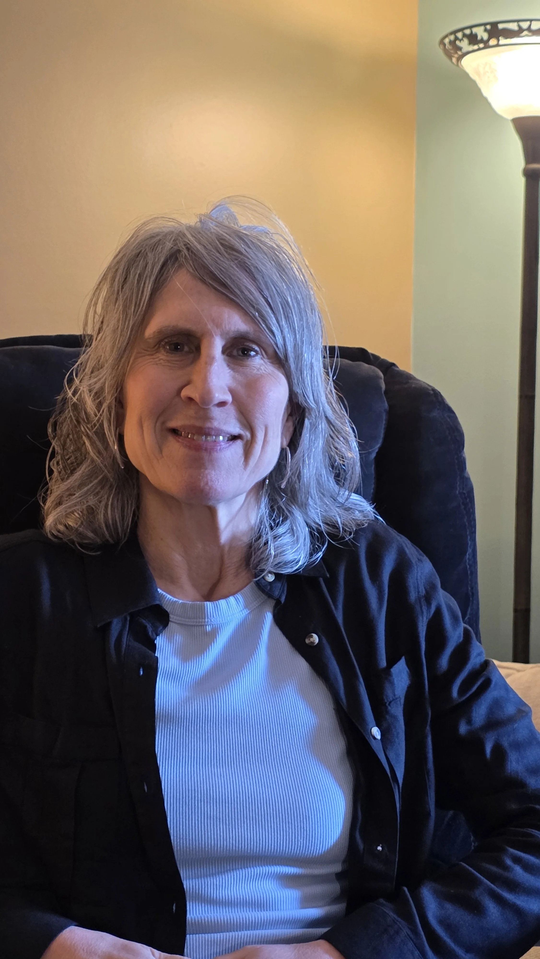 A smiling middle-aged woman with gray hair sitting on a black chair in a room with yellow and green walls, next to a floor lamp.