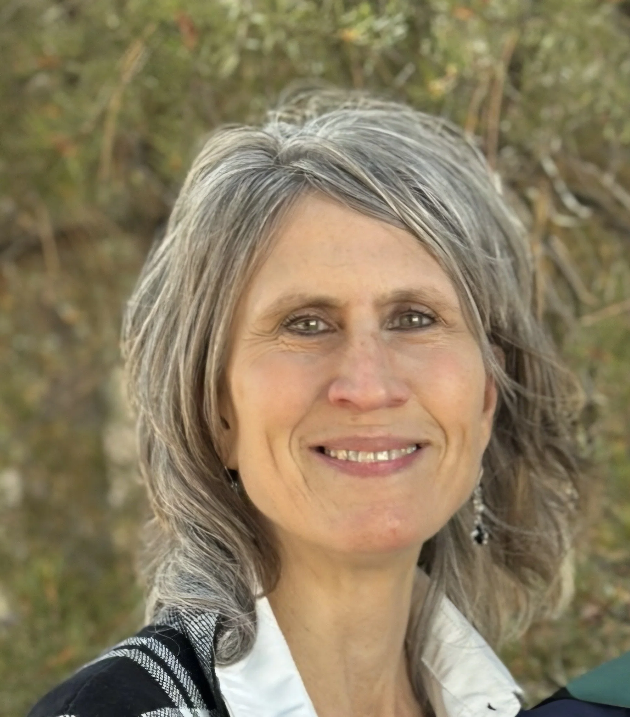 Close-up of a smiling woman with gray and black hair, outdoor background with green foliage.