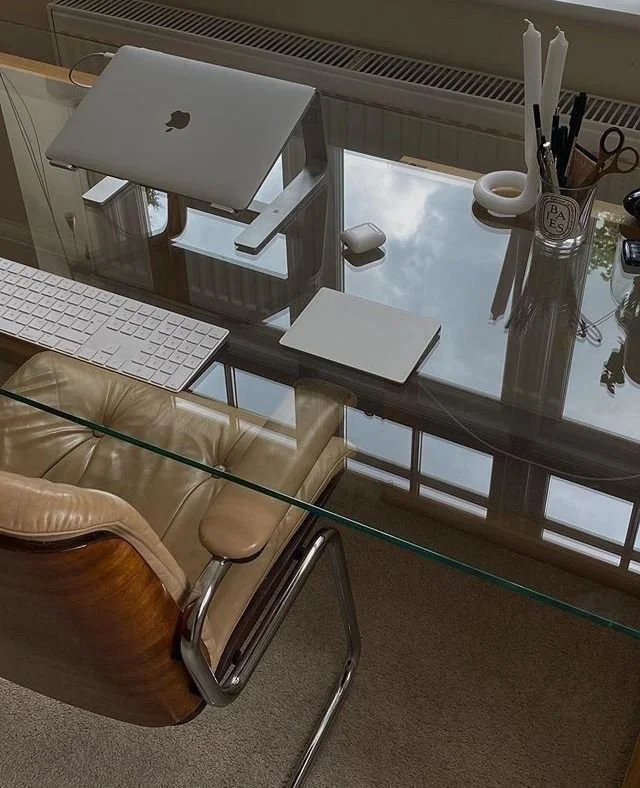 A modern workspace with a glass desk, an Apple MacBook, a wireless keyboard, a notepad, and office supplies. A beige leather chair is pushed in at the desk, and a window with a radiator is in the background.