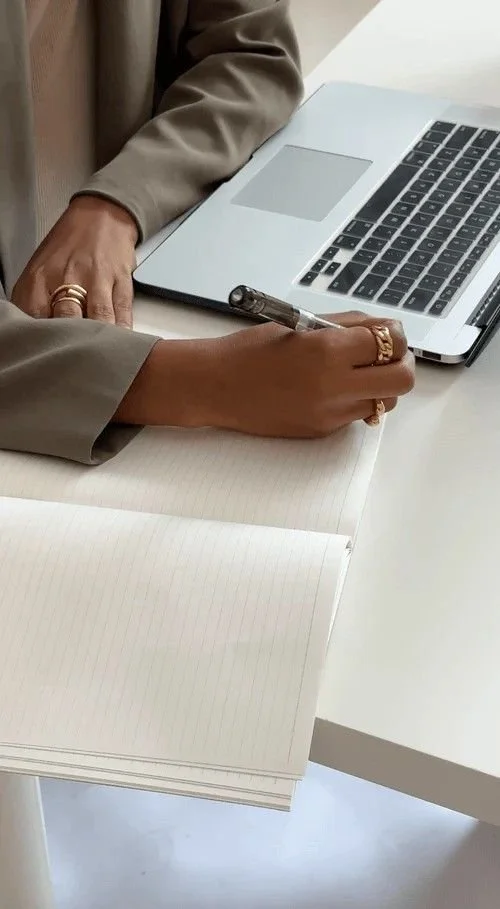 Person writing in an open notebook with a pen, beside a laptop on a white desk.