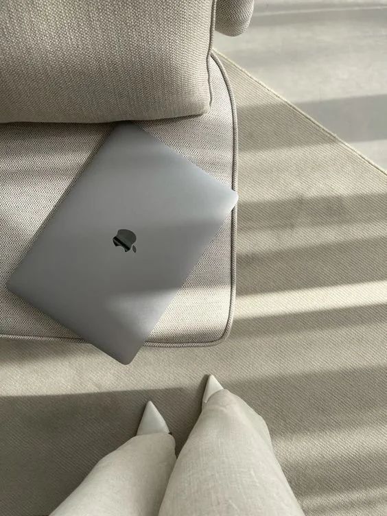 A closed silver MacBook laptop resting on a light-colored sofa with sunlight streaming in.