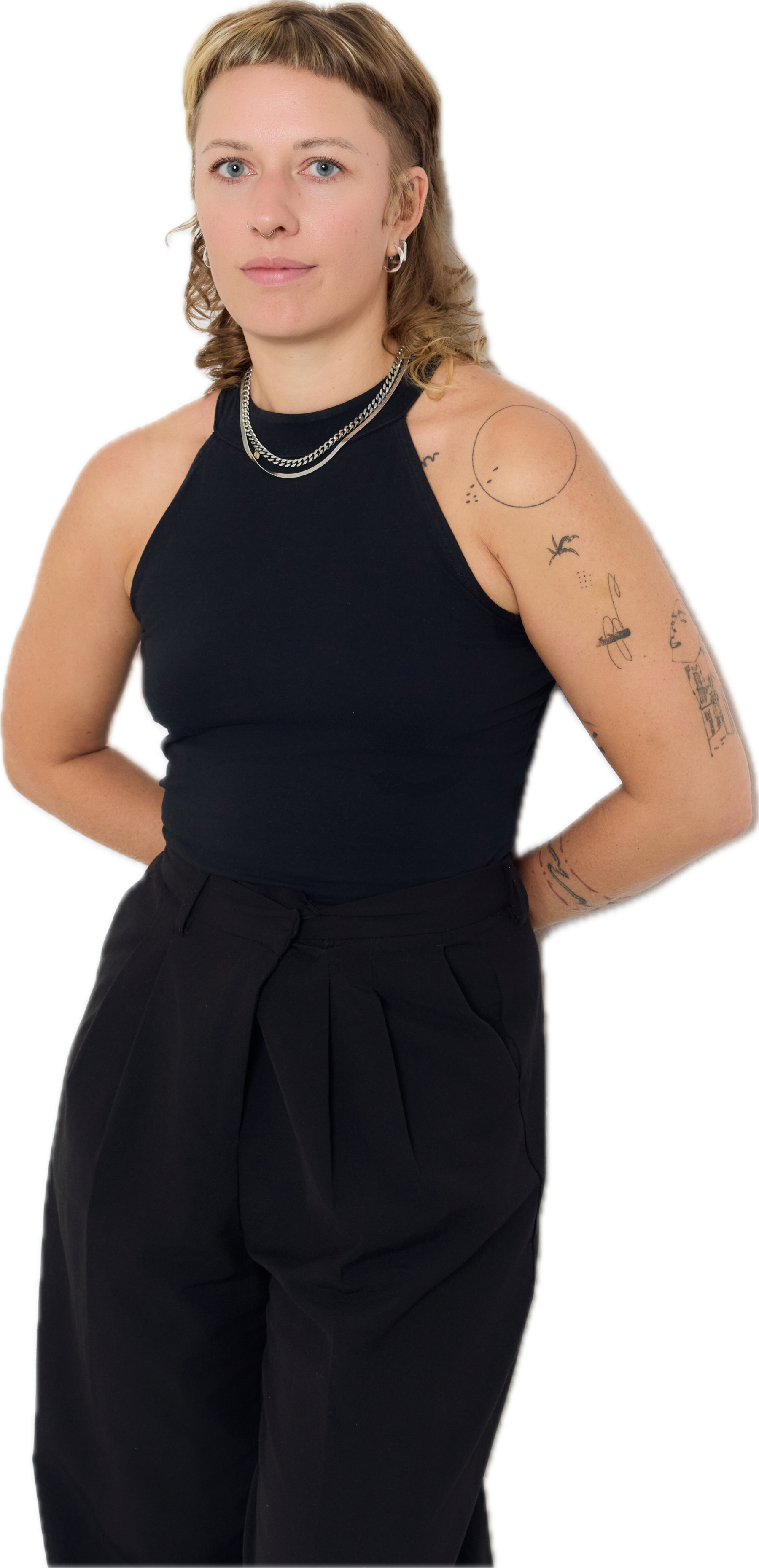 Junge Cis-Frau mit kurzen, zweifarbigen Haaren, trägt schwarzes Top und Hose, steht mit hinter dem Rücken verschränkten Armen, trägt Halsketten und hat Tattoos am Arm und Schulter.
