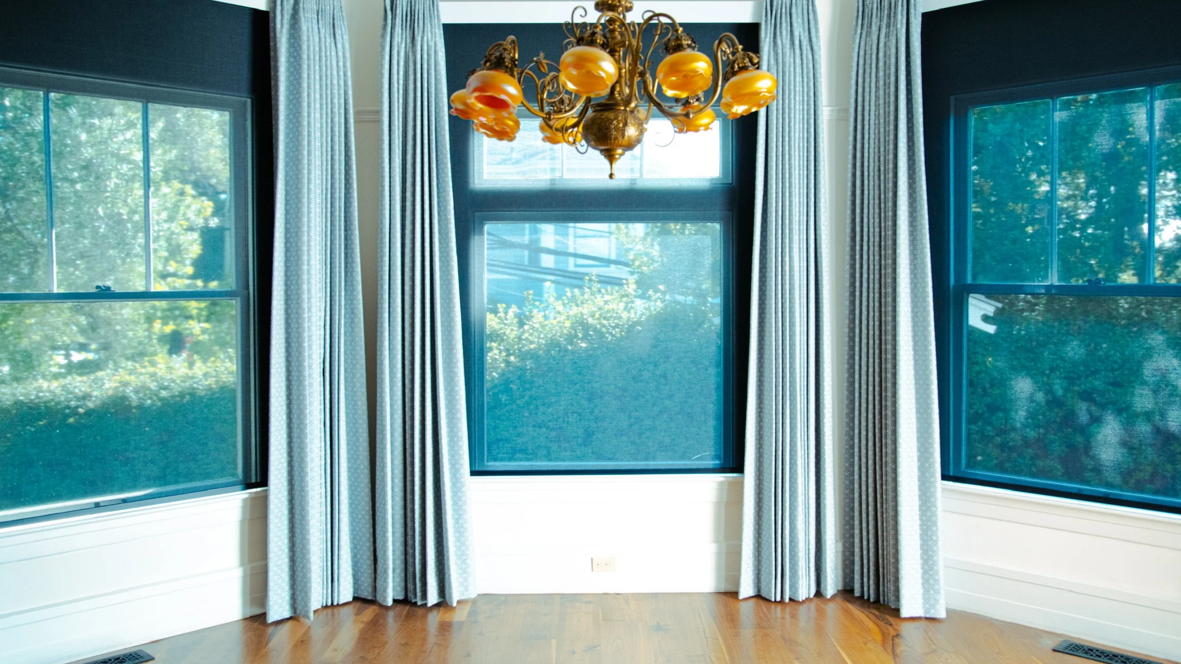 A room with large windows, light blue curtains, and a vintage chandelier with orange glass shades hanging from the ceiling.
