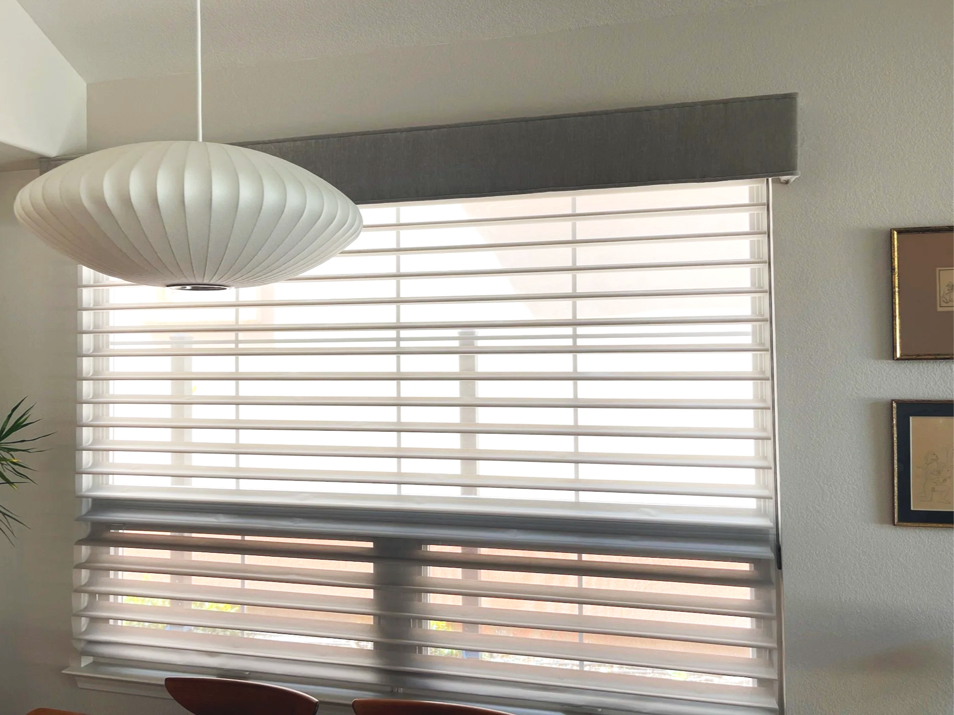 A window with white horizontal blinds partially open, with light coming through. A white paper pendant light hangs from the ceiling in the foreground. On the right side of the image, part of a wall with a framed picture is visible.