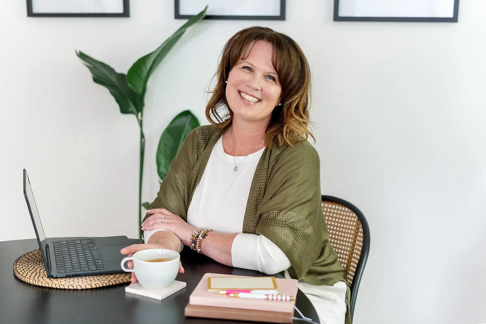 A smiling woman with short brown hair wearing a white top and an olive green shawl, sitting at a desk with a laptop, a cup of tea, a pink planner, and pens, in a bright room with a large green plant behind her.