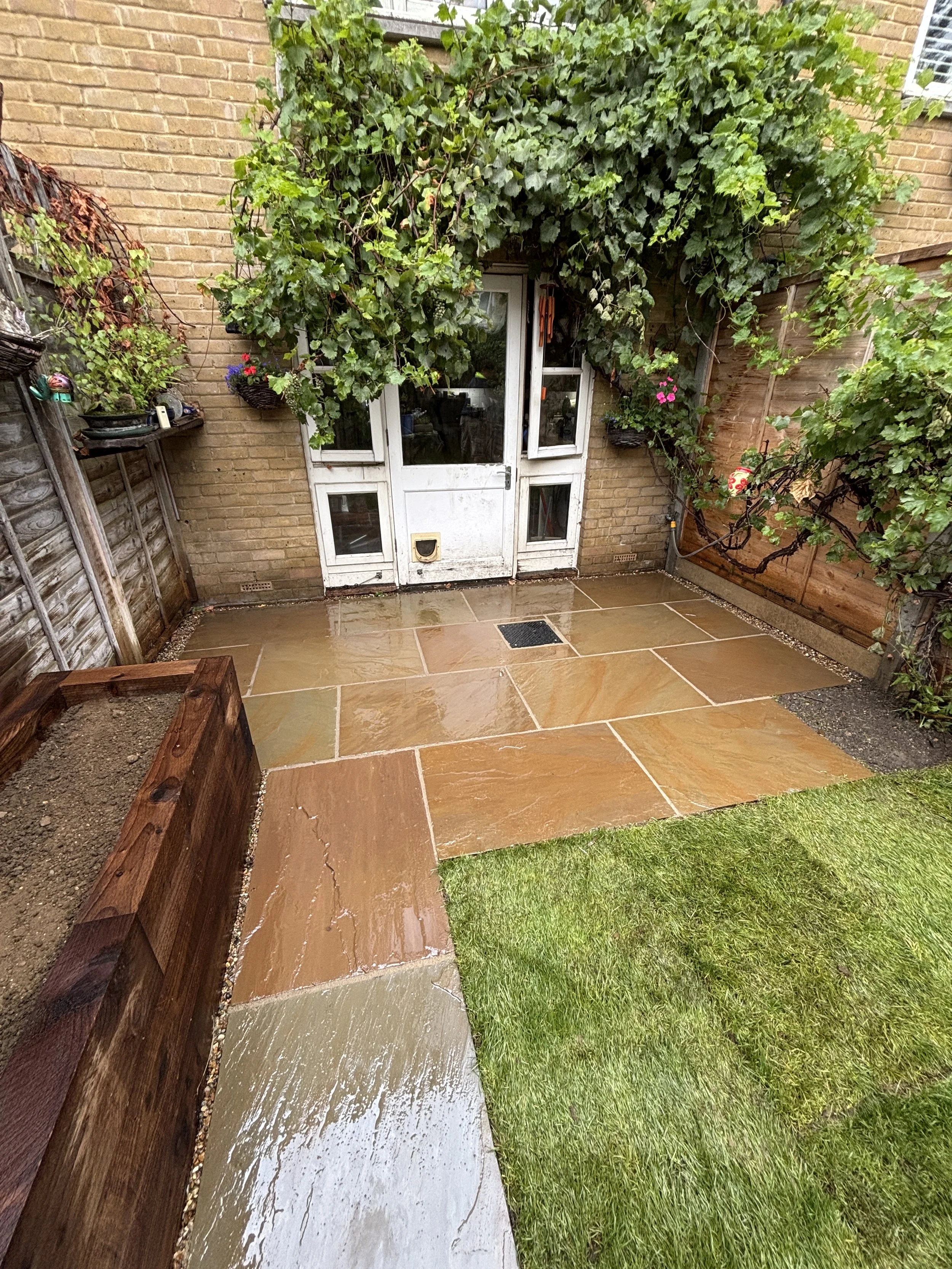Wet outdoor patio with stone tiles, a small patch of grass, potted plants, a brick wall with green ivy, and a white door with glass panels.Garden design near me, East london garden design. West london garden design. North London Garden design.