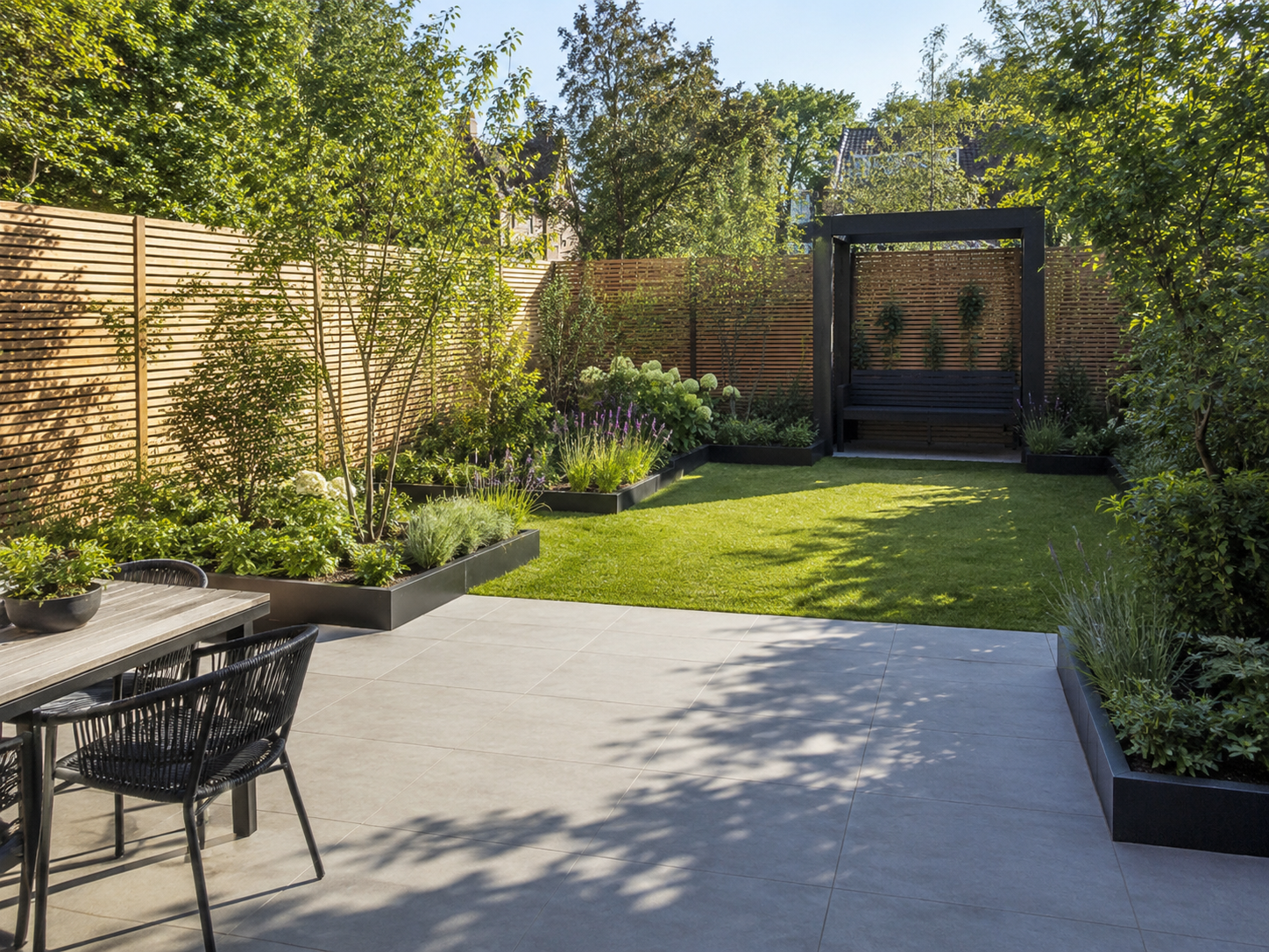 A backyard garden with a patio, black outdoor furniture, lush green grass, flower beds with purple and white flowers, wooden privacy fence, and a black garden seat within a black frame structure.