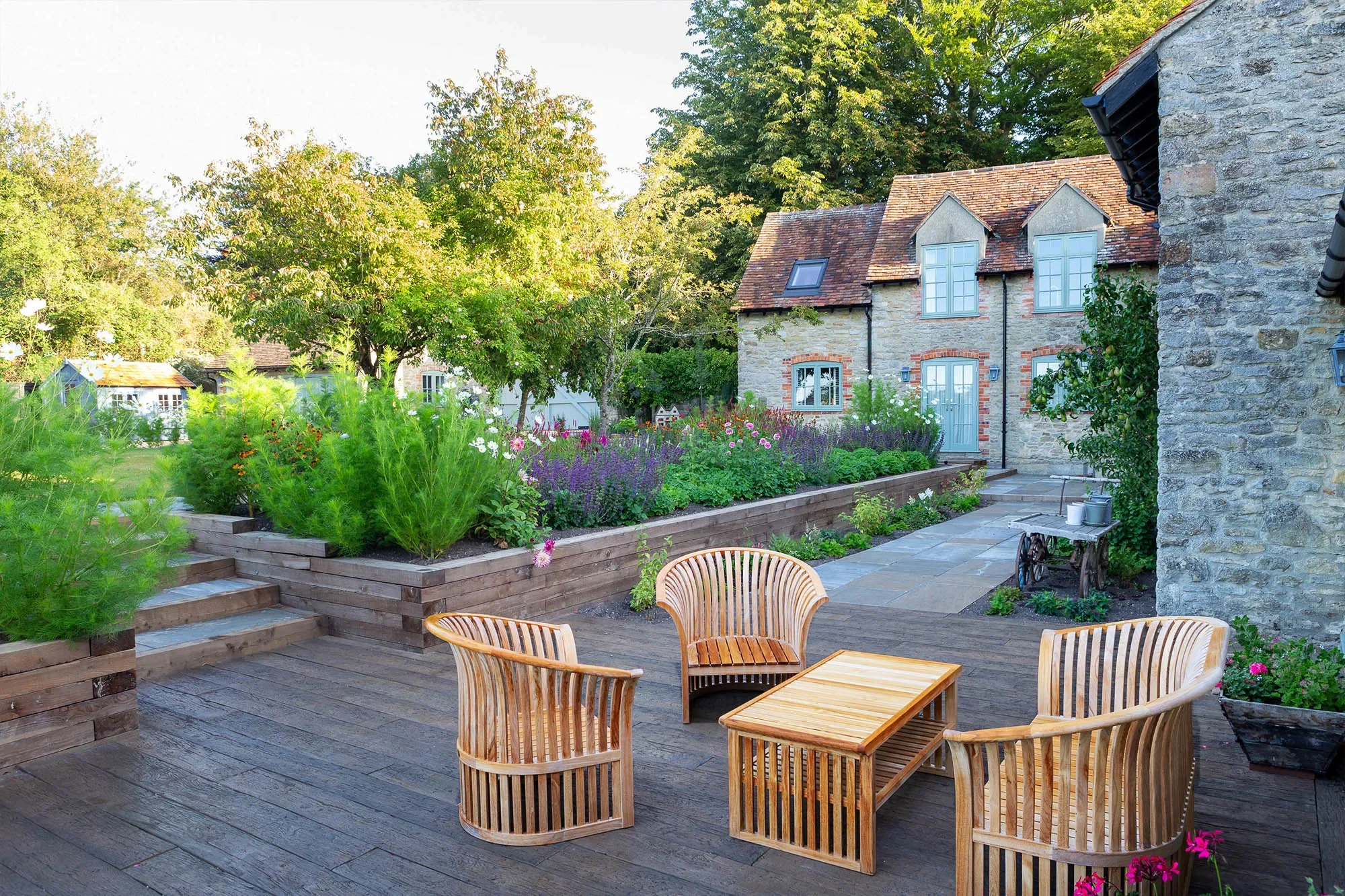 A quaint backyard patio with wooden outdoor furniture, including three chairs and a small table, surrounded by green plants and flowers, with a stone house and mature trees in the background. Garden design near me, East london garden design. West lon
