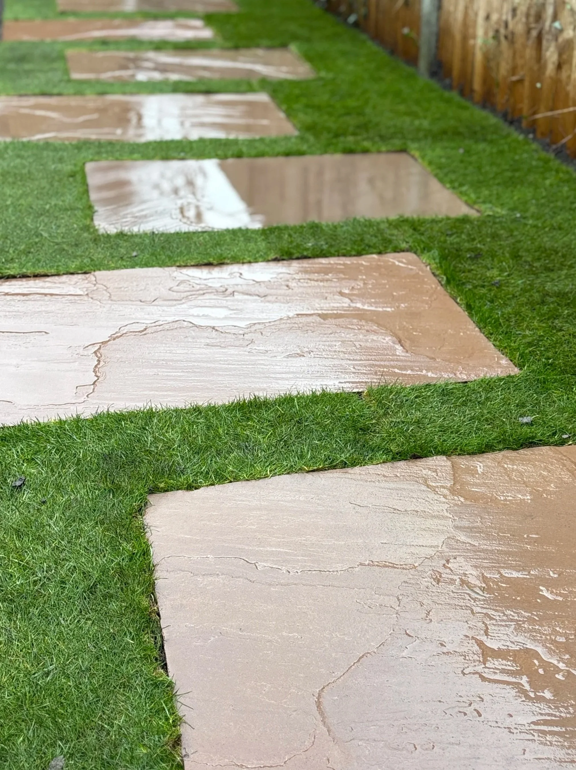 Close-up of a pathway with rectangular stone slabs and grass in between, and a wooden fence in the background. Garden design near me, East london garden design. West london garden design. North London Garden design.