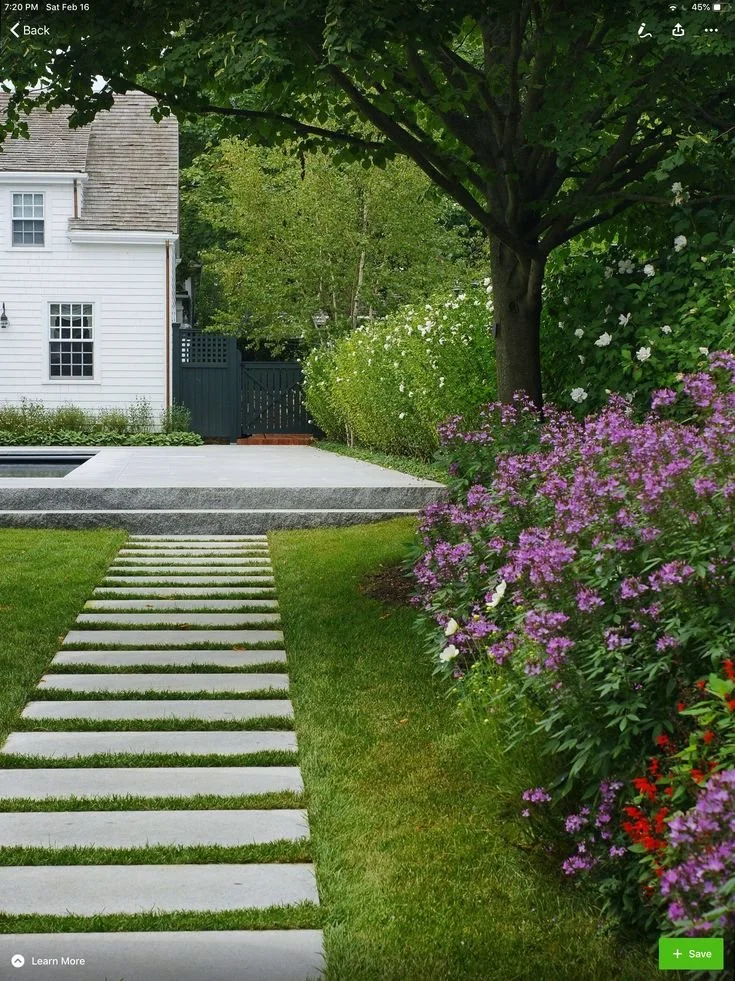 A backyard garden with a stone pathway, green grass, flowering bushes, a large tree, and a white house with a dark gate in the background.London garden design.