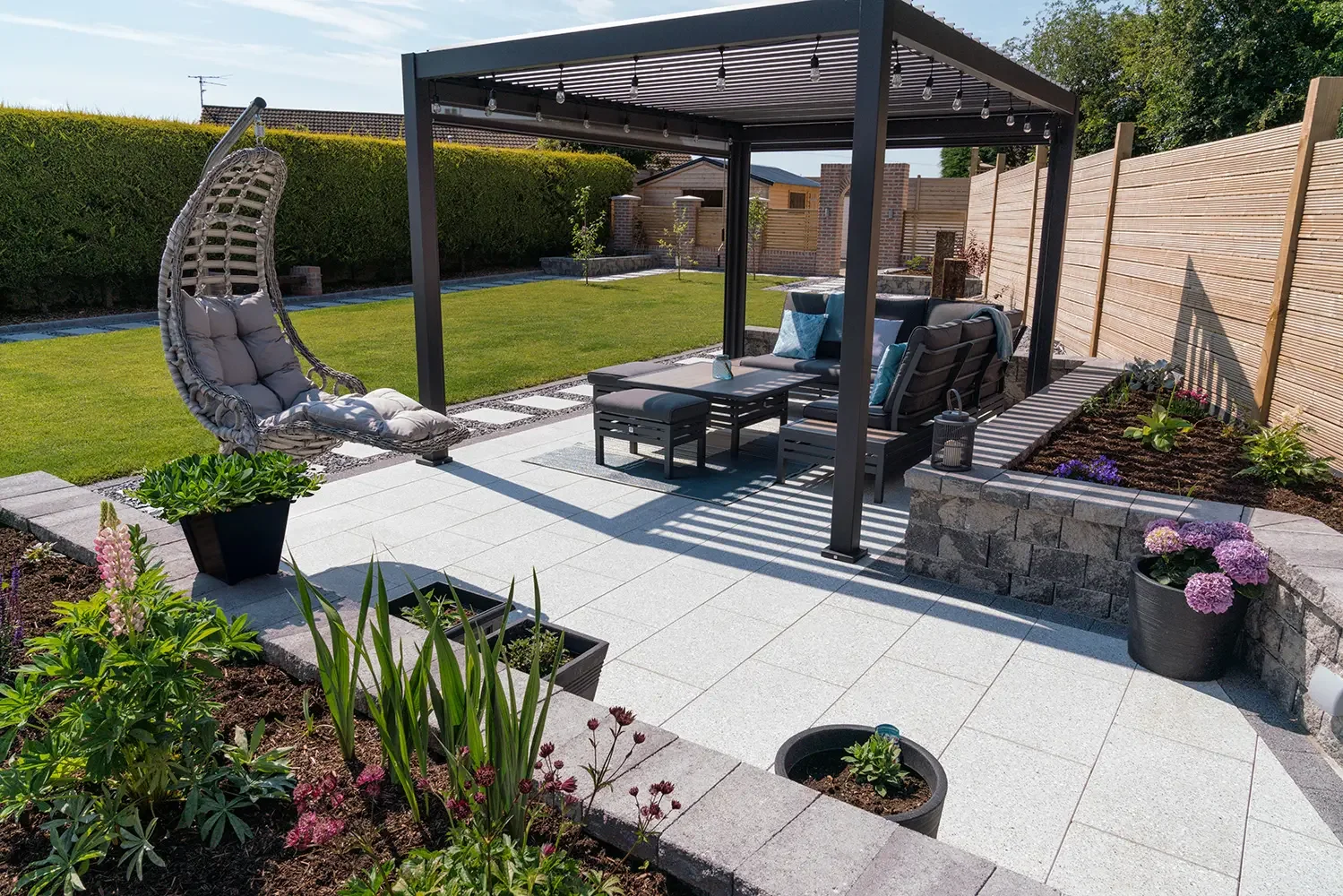 A backyard patio with outdoor seating, a hanging chair with cushions, potted plants, a covered pergola with string lights, a tidy lawn, and a wooden fence and garden beds along the sides. Garden design near me, East london garden design. West london