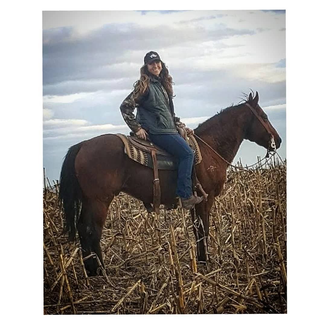 A woman sitting on a brown horse in a field of dry corn stalks, wearing a cap, jacket, jeans, and smiling.