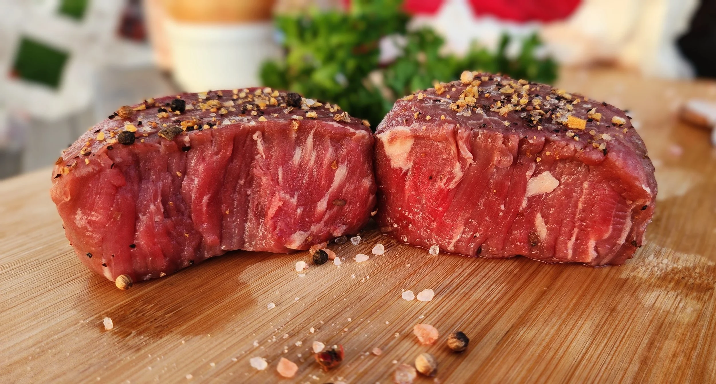 Two thick cuts of raw beef seasoned with salt and pepper on a wooden cutting board.