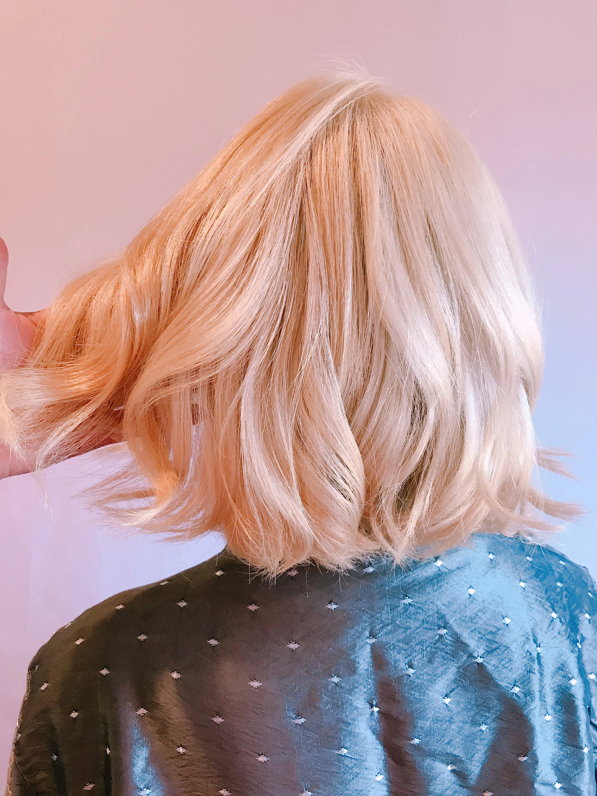 A person with shoulder-length blonde hair styled in loose waves, seen from behind, wearing a black shirt with small white patterns.