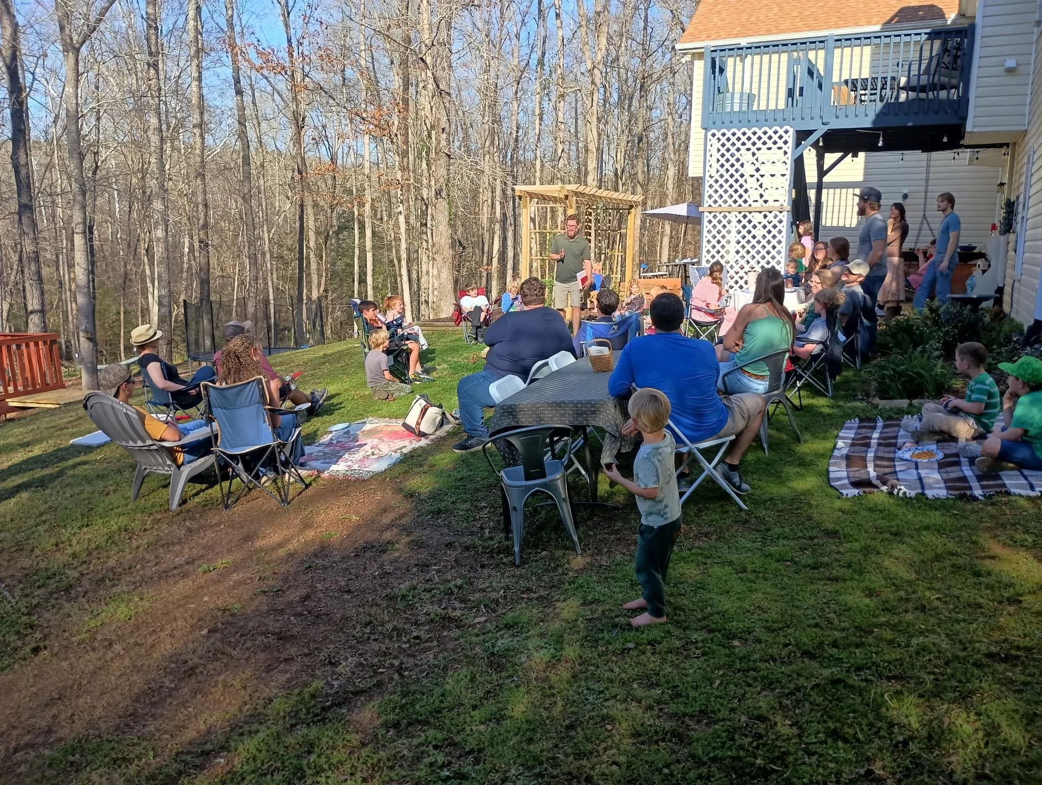 A backyard gathering with people seated on lawn chairs and blankets, listening to a man speaking in front of trees on a sunny day.