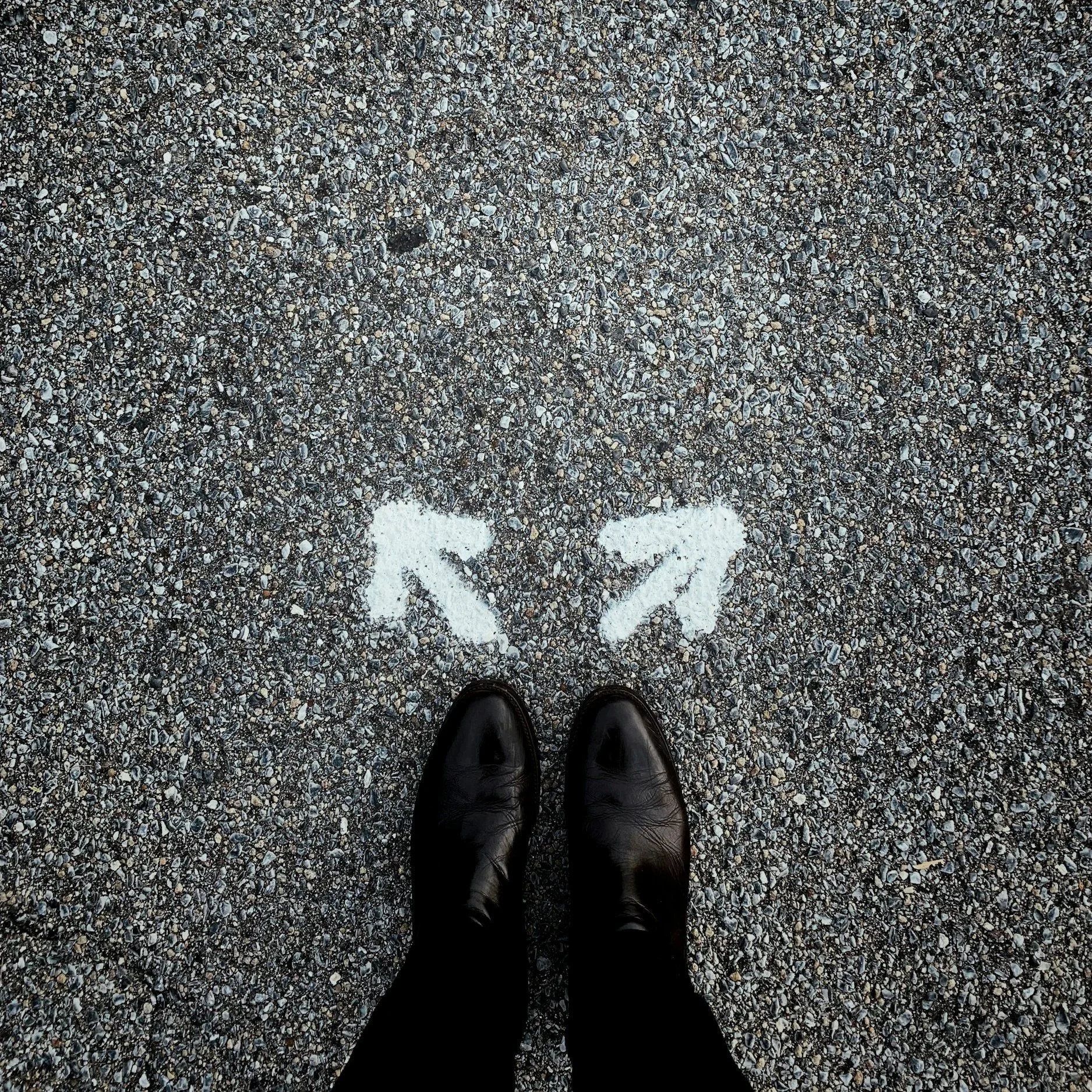 A person's black shoes standing on gravel with an arrow on the ground pointing to the left.