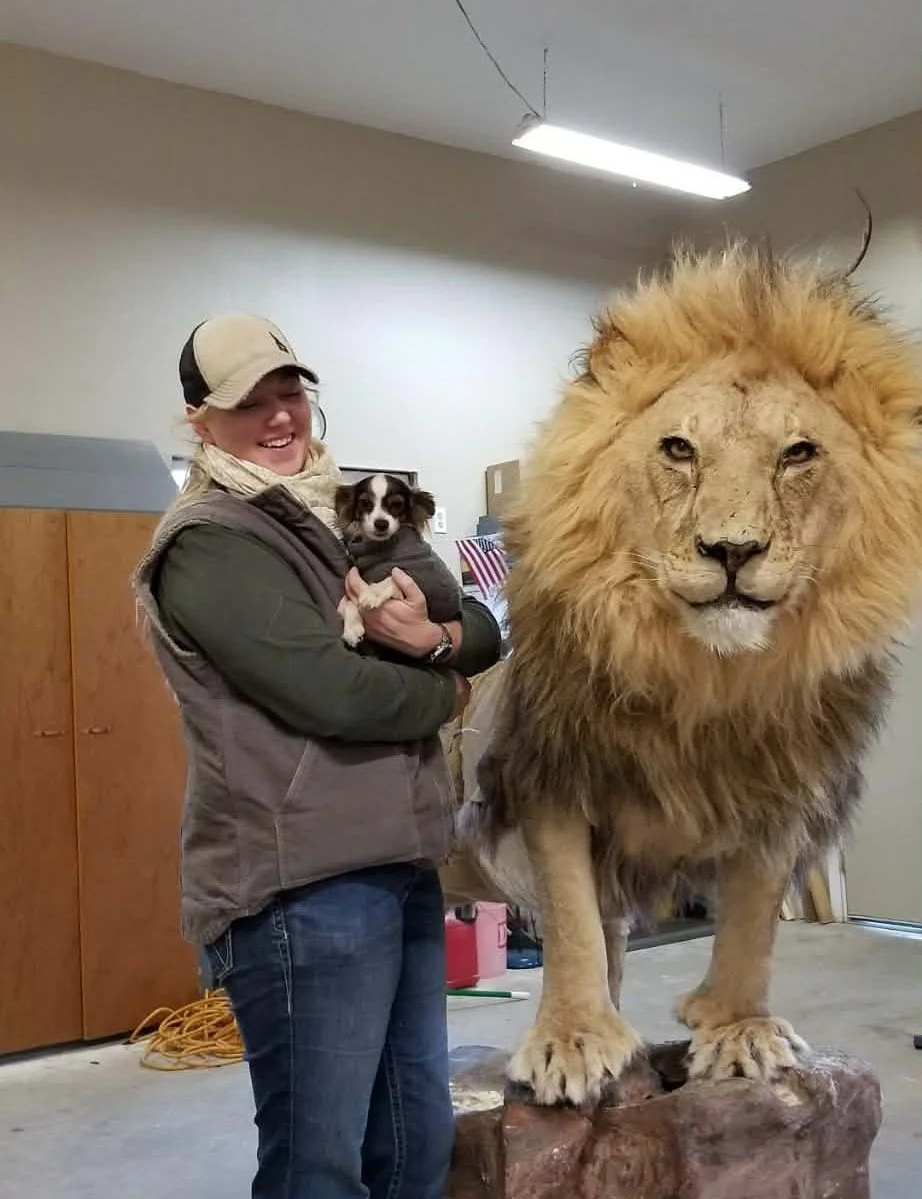 A woman smiling while holding a small dog in front of a large taxidermy lion in an indoor setting.