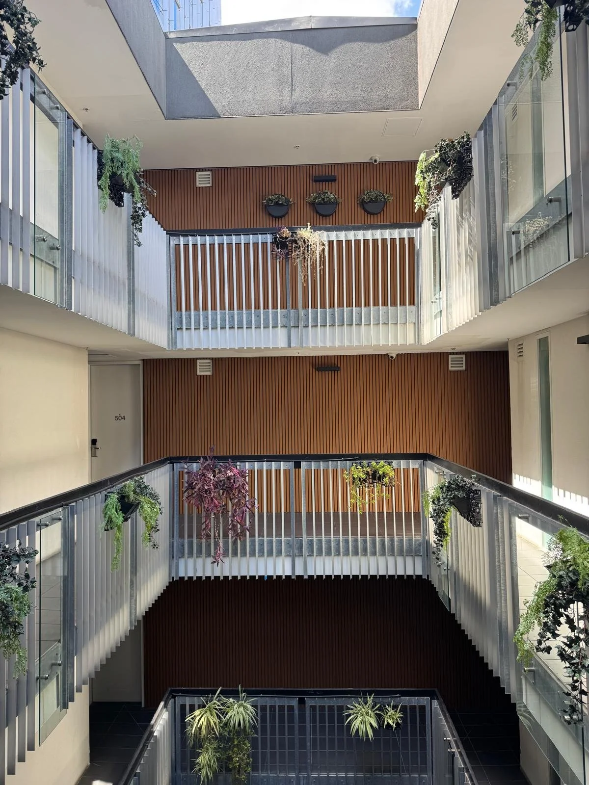 Multiple floors of a modern building with white railings, wooden wall panels, and potted plants including hanging and ground plants, arranged along the corridors.
