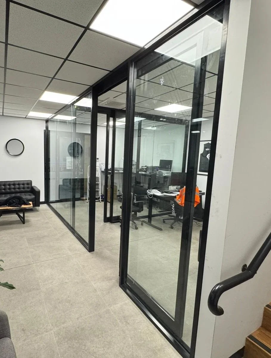 An office with glass partition doors, a waiting area with a black couch, and a desk with a chair and office supplies.