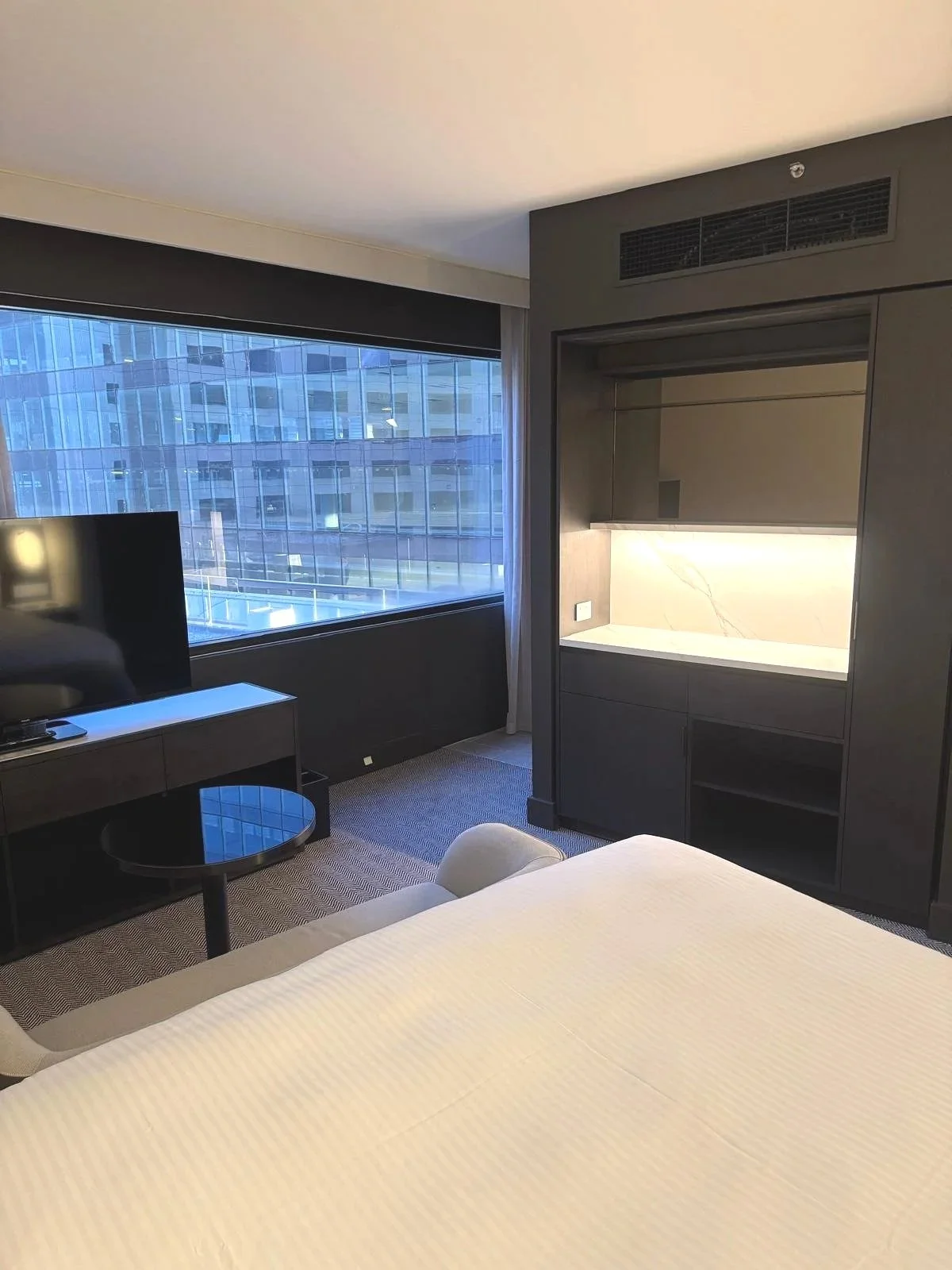 Hotel room with a large window, a flat-screen TV on a stand, a small round black table, a white bed, and a built-in cabinet with a marble backsplash.