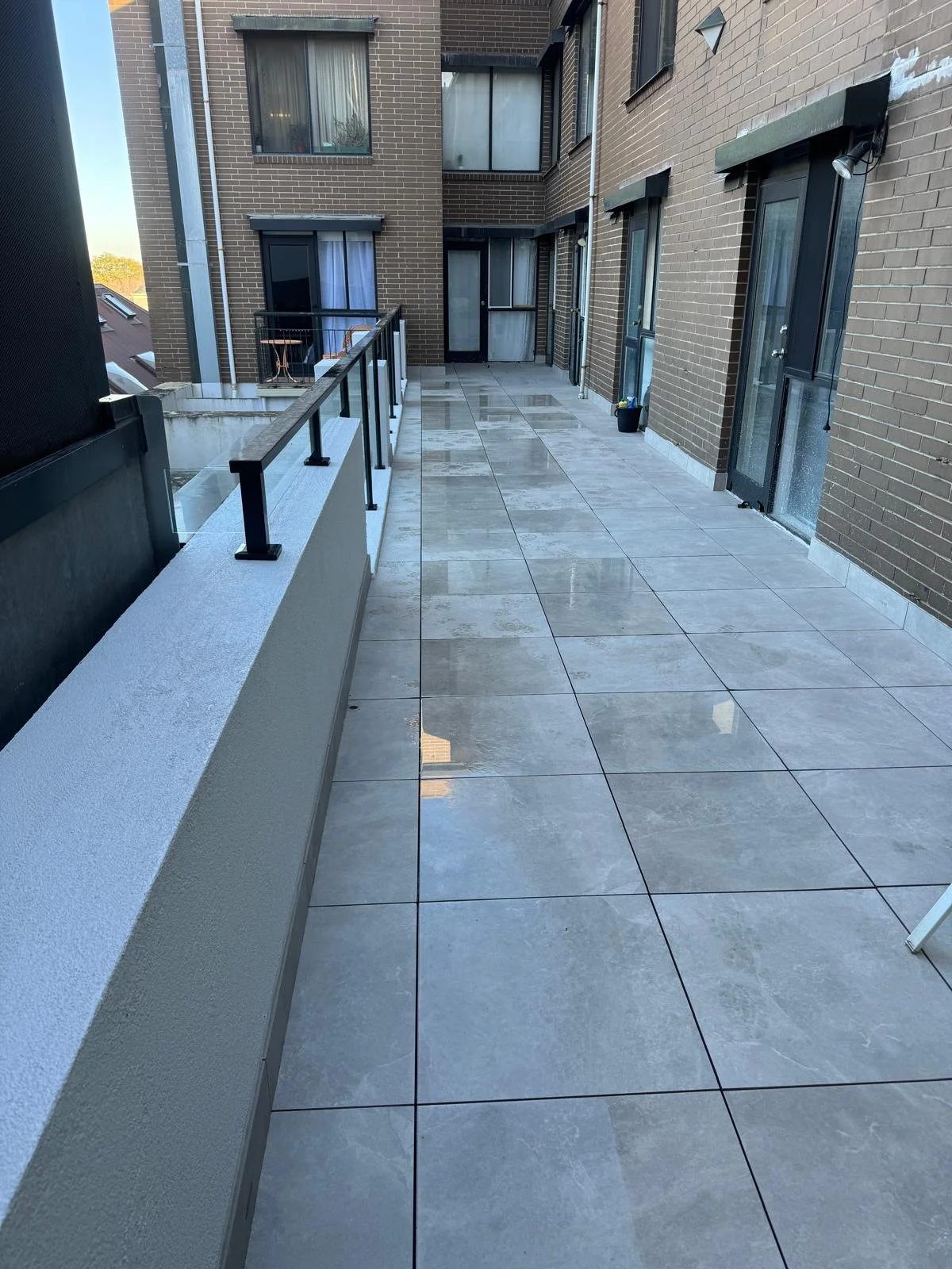 Empty balcony with wet tile floor, brick apartment building walls, black metal railing, and glass doors and windows.