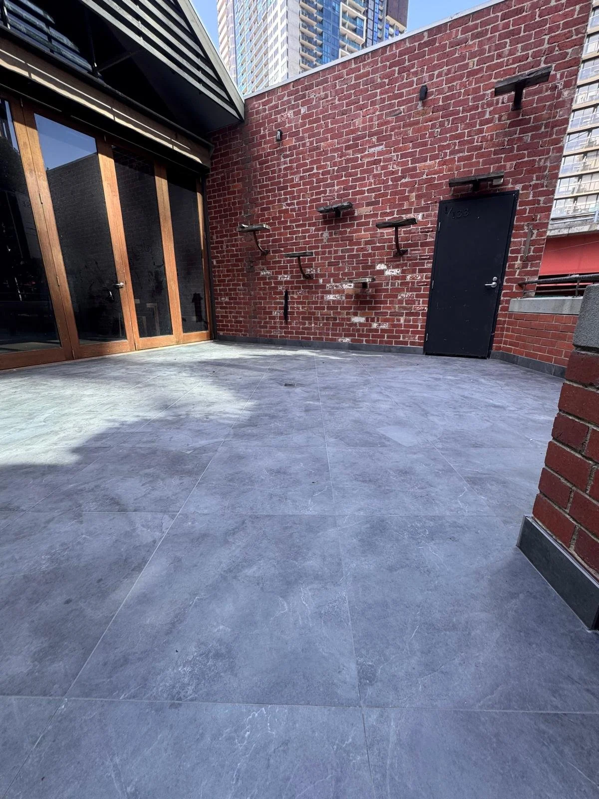 Empty outdoor balcony with gray tiled floor, red brick walls, and a black door, with city buildings in the background.