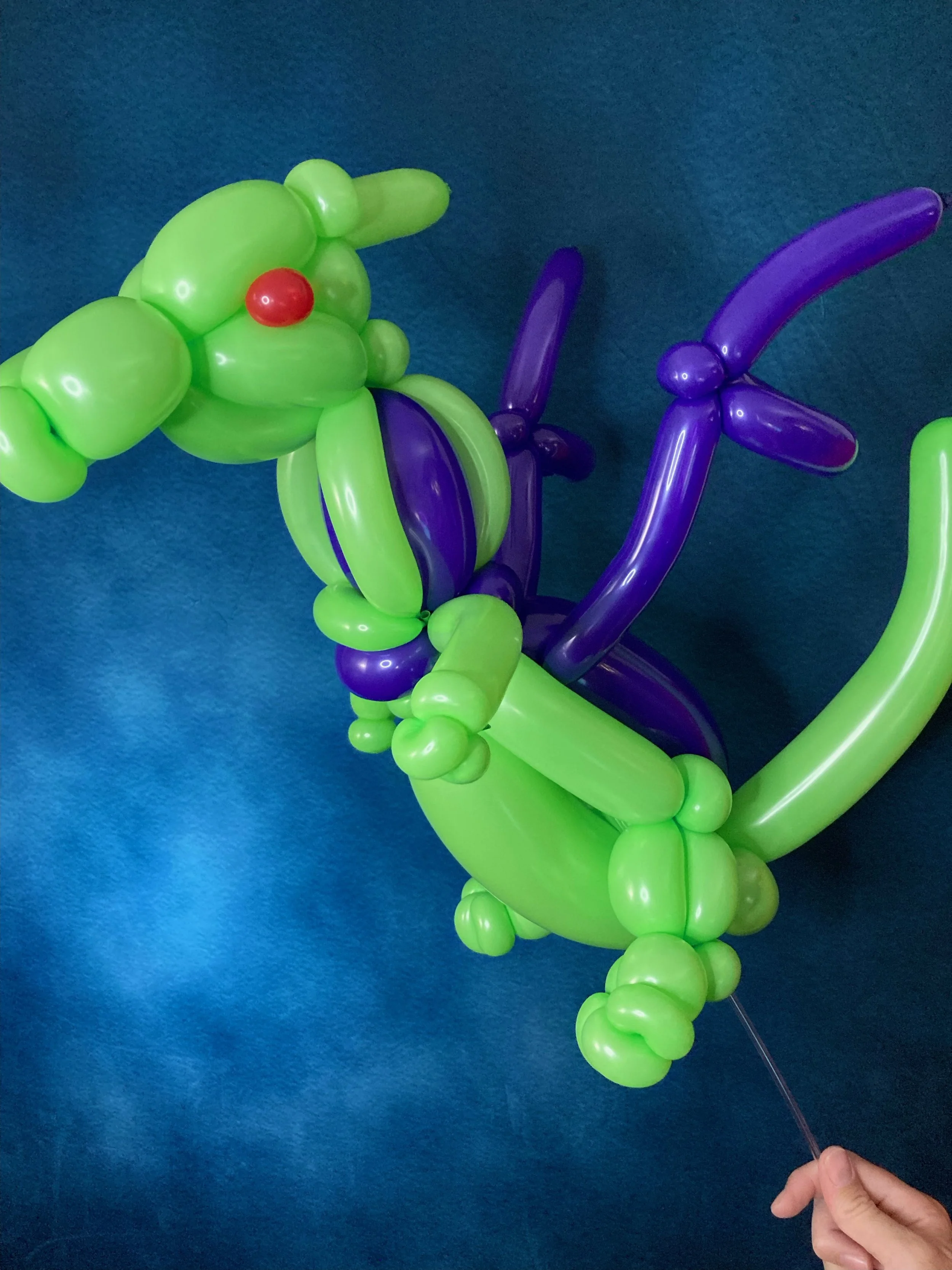 Balloon art sculpture resembling a dinosaur, primarily green with purple and red accents, held by a person's hand.