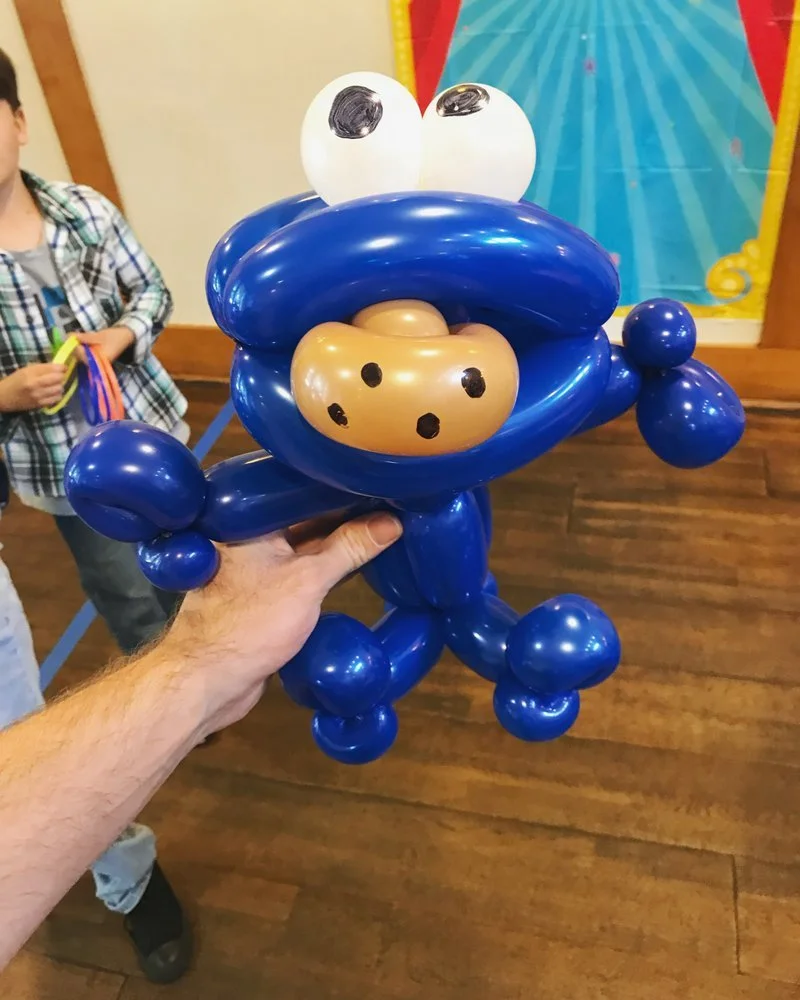 Balloon sculpture resembling a blue cartoon character with large white eyes and black pupils, a beige nose with black dots, and a large open mouth.