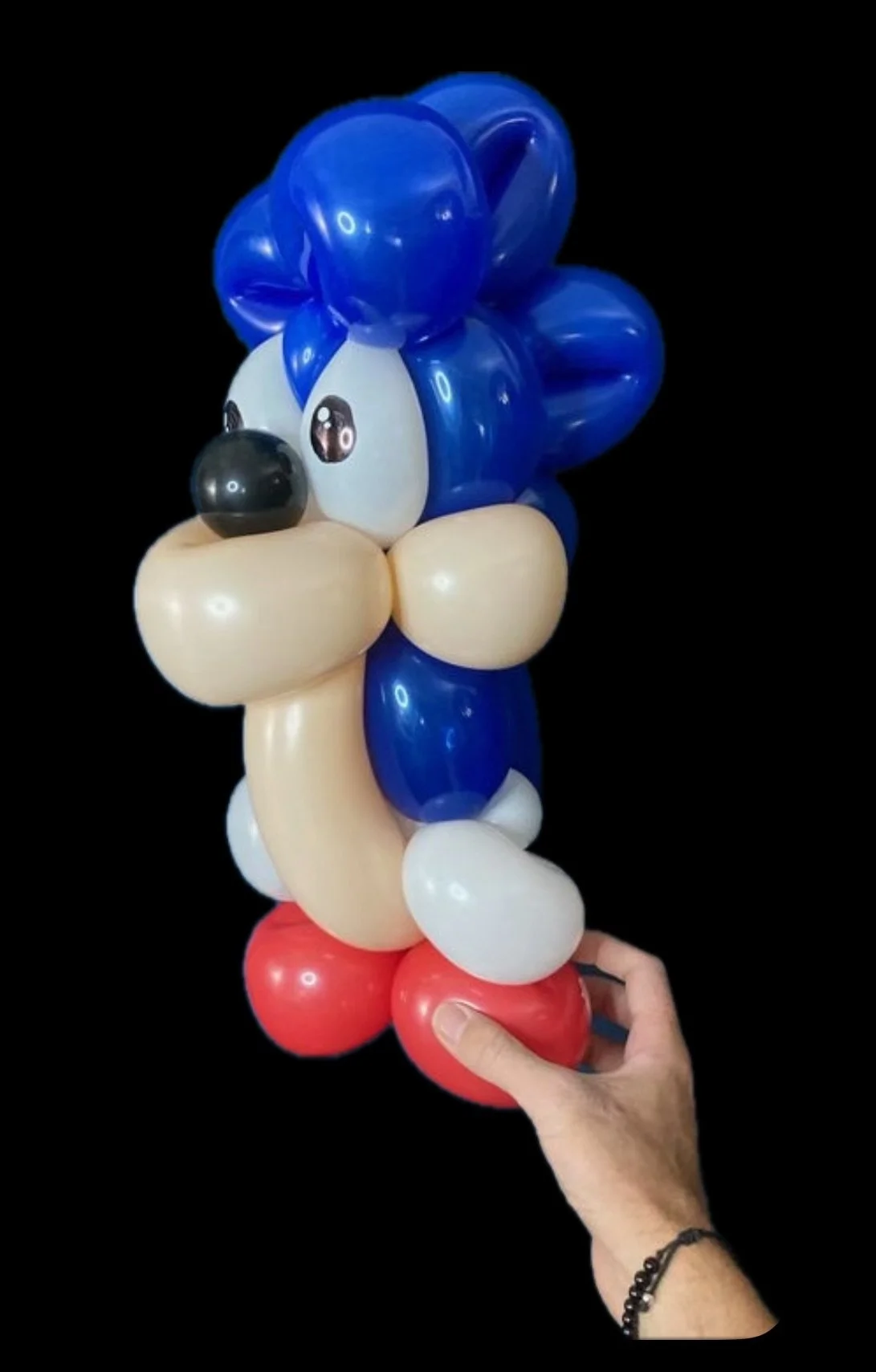 Balloon sculpture of Sonic the Hedgehog with blue hair, white gloves, red shoes, and a beige body, held by a person against a black background.