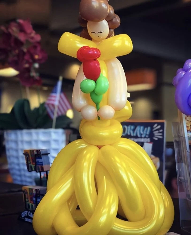 Balloon sculpture of a young girl with brown hair, yellow dress, holding a bunch of red and green balloons.