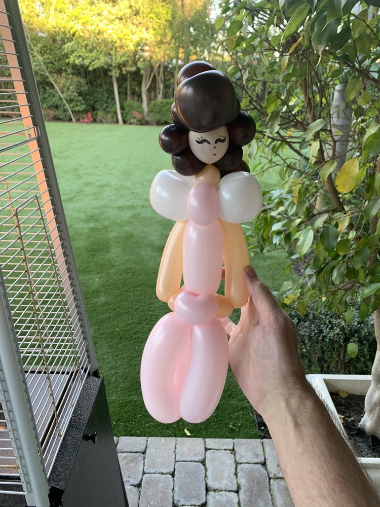 Balloon sculpture of a girl with black hair, rosy cheeks, wearing pink and cream-colored clothing, held by a person's hand outdoors with greenery in the background.