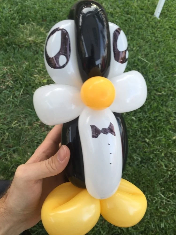 Balloon sculpture of a dog with black and white colors, a bow tie, and yellow balloon feet, held in a person's hand outdoors on grass.