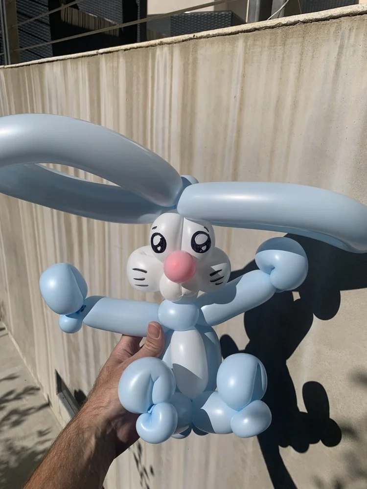 Balloon sculpture of a cartoon bunny with long ears, pink nose, and expressive eyes, being held outdoors.