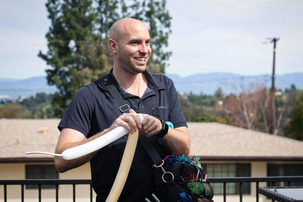 A smiling man with a shaved head standing outdoors, holding white and tan balloons, with a residential area and mountains in the background.