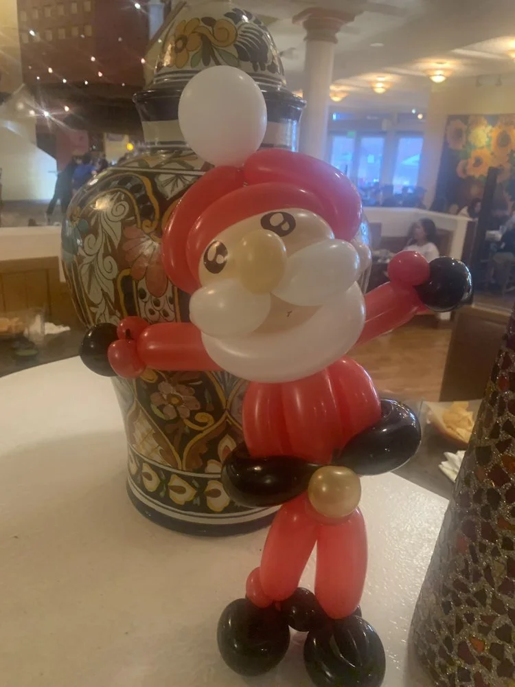 Balloon sculpture of Santa Claus with a red hat, white beard, black shoes, and red outfit, standing on a table at a festive indoor event.