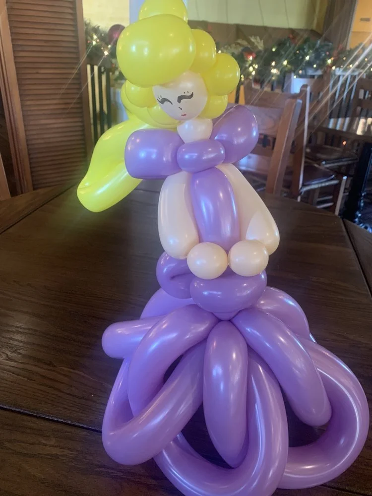 Balloon sculpture of a girl with long yellow hair, purple dress, and a purple flower-shaped skirt, placed on a wooden table