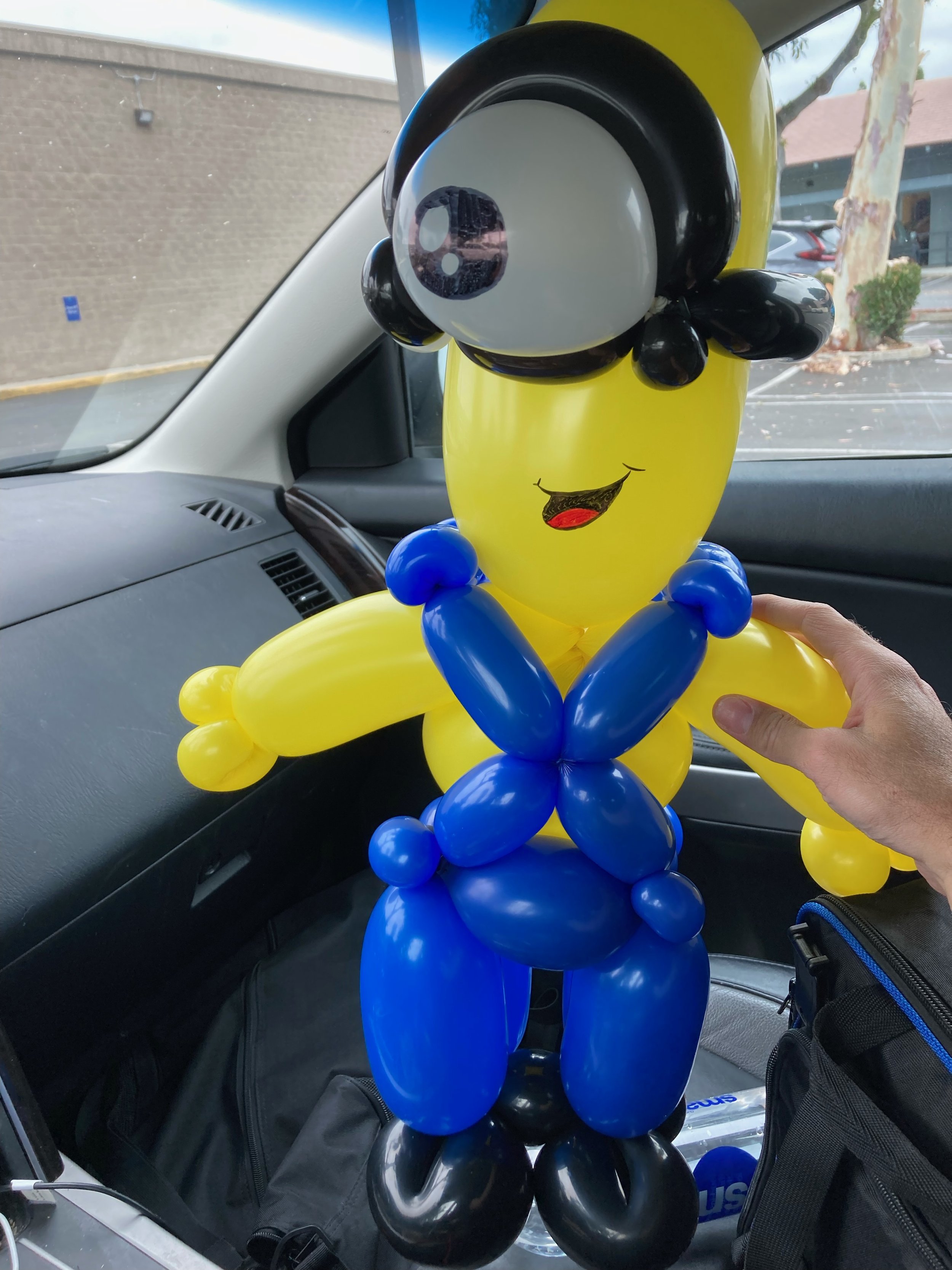 Balloon sculpture of a cartoon character with large eyes, yellow face and body, black hair, wearing blue shorts and gloves, sitting in a car.
