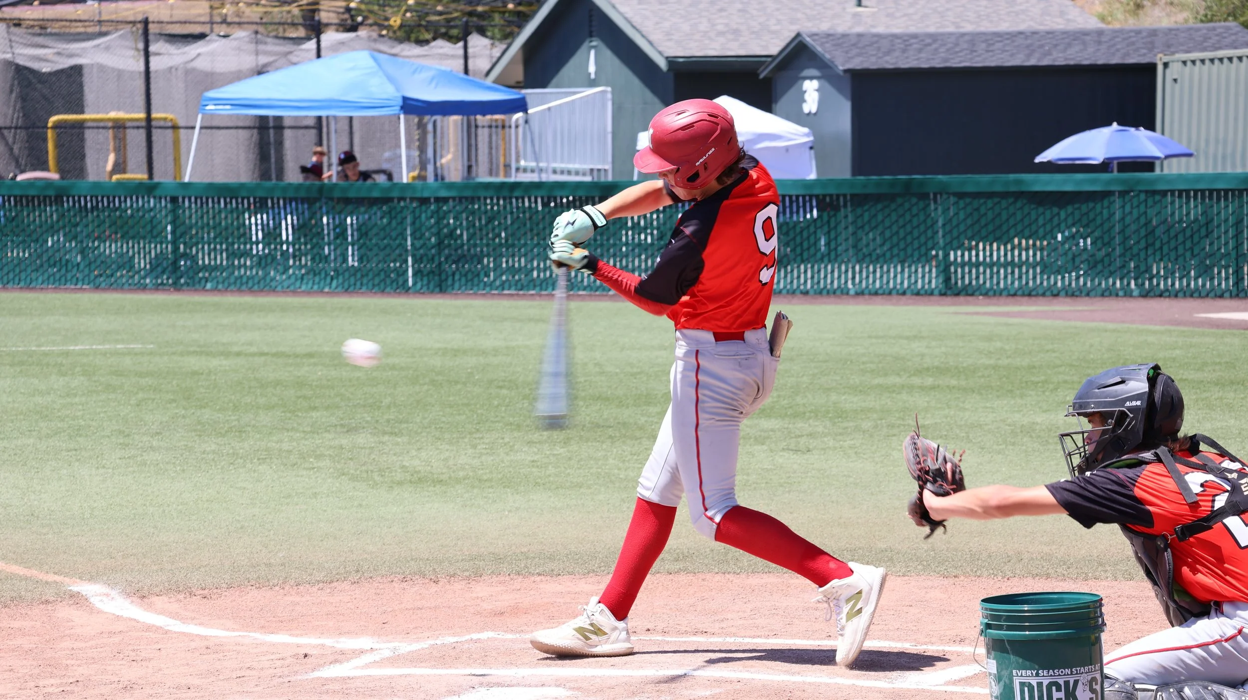 A young baseball player in a red helmet and uniform is hitting a pitched ball with a bat during a game. A catcher in a black and red uniform and helmet is in a crouched position behind home plate, reaching out with a glove. The field has a dirt infie