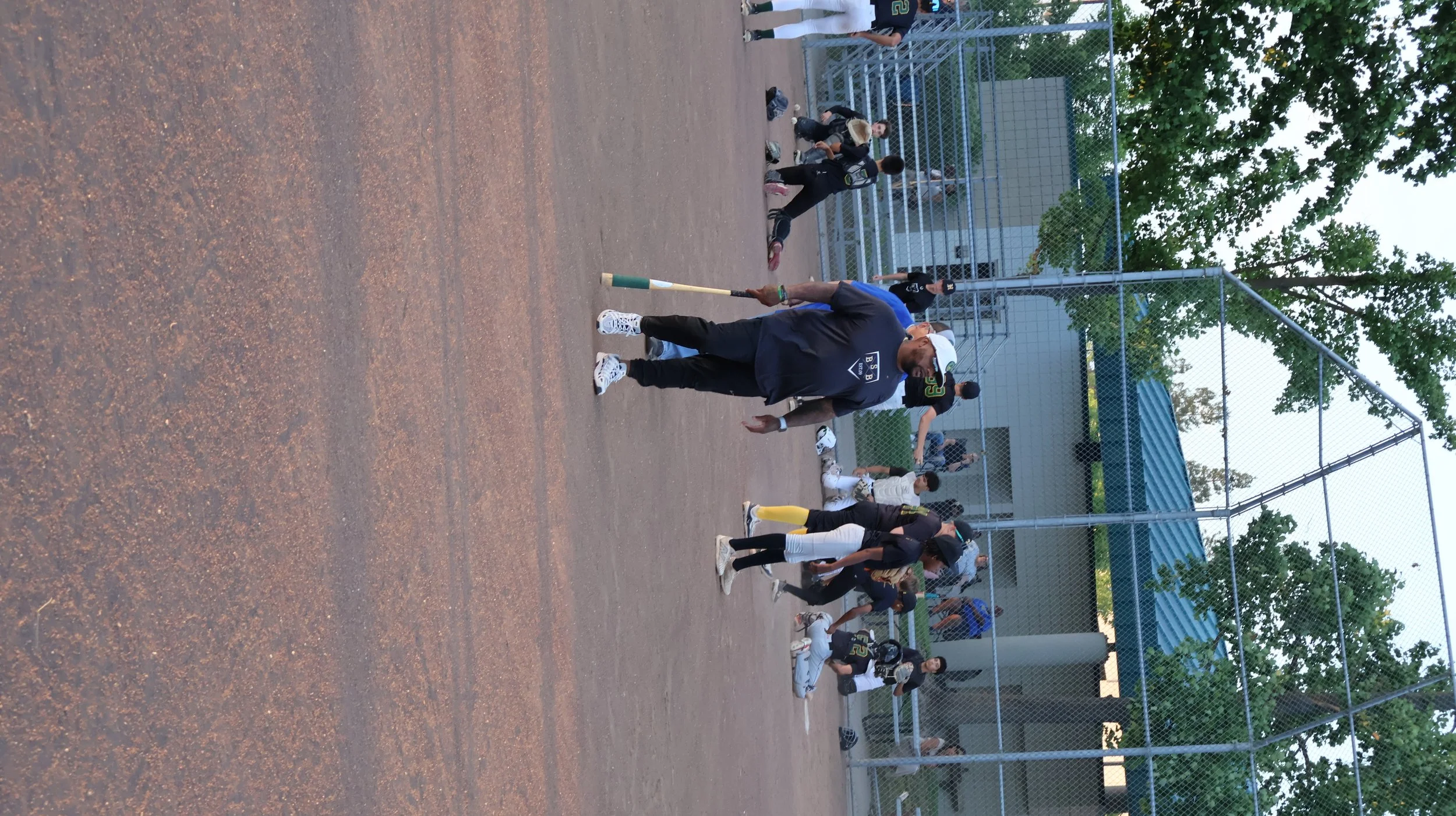A baseball coach and team on the field of a baseball diamond during a game, with players sitting and standing near the dugout and spectators in the background.