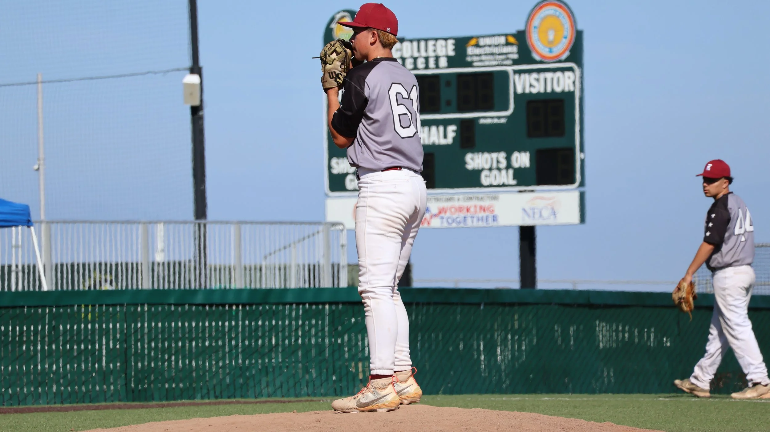 Two young baseball players on the field, one standing on the pitcher's mound and the other walking with a glove, during a game in bright weather.