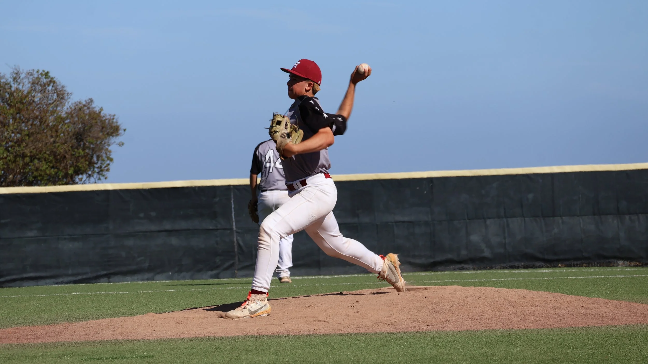 A young baseball player in a black and gray uniform with a red cap is pitching on the mound during a game, with another player visible in the background.