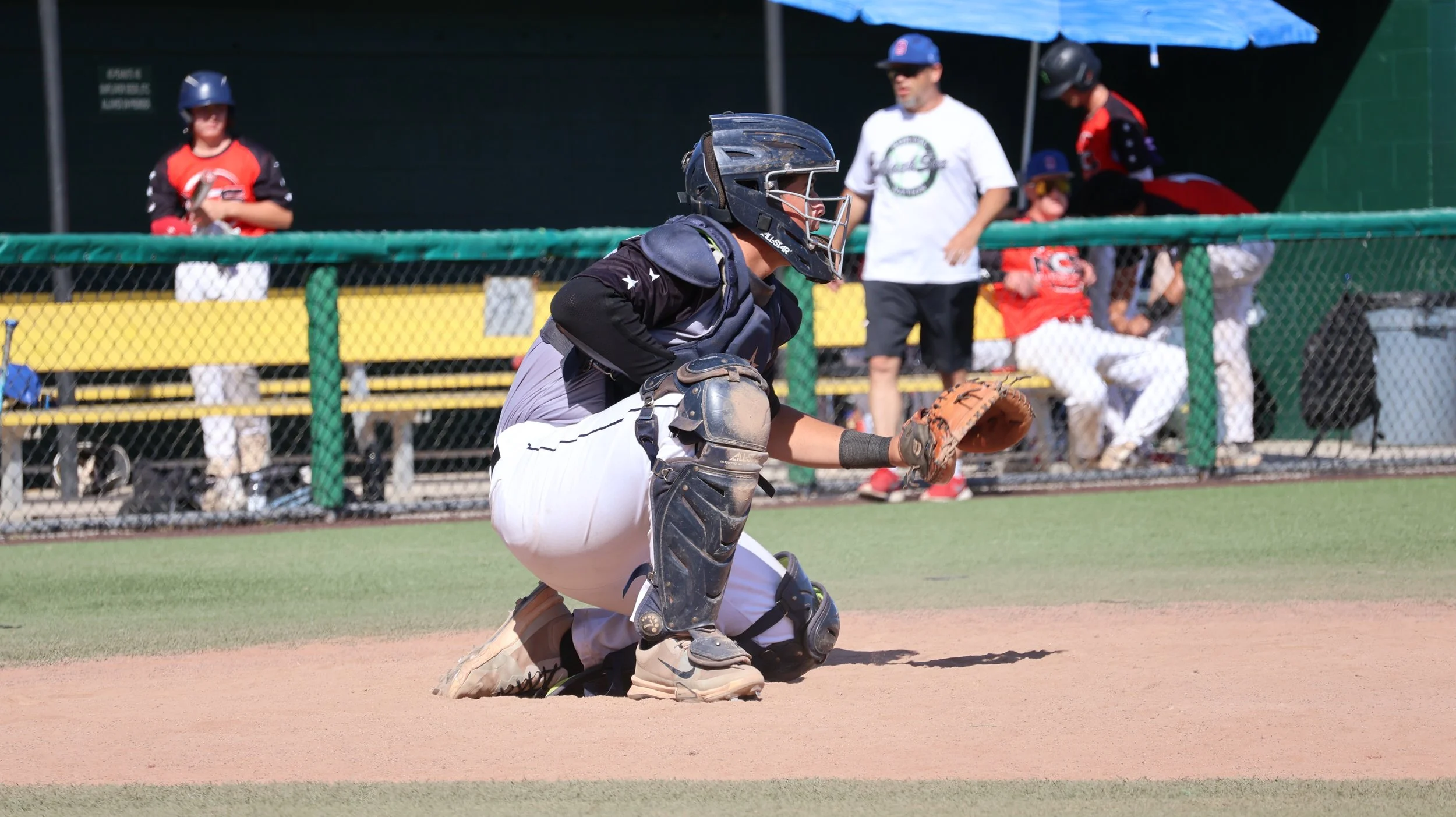 A baseball catcher in protective gear crouching on the field during a game, with spectators and team members sitting and standing behind a green fence and under a blue umbrella.