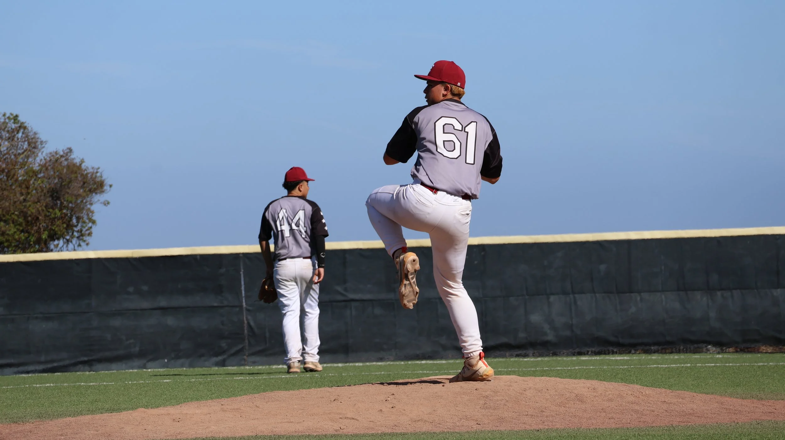 Two baseball players in uniforms and red caps on a baseball field, one pitching and the other standing, with a blue sky and trees in the background.